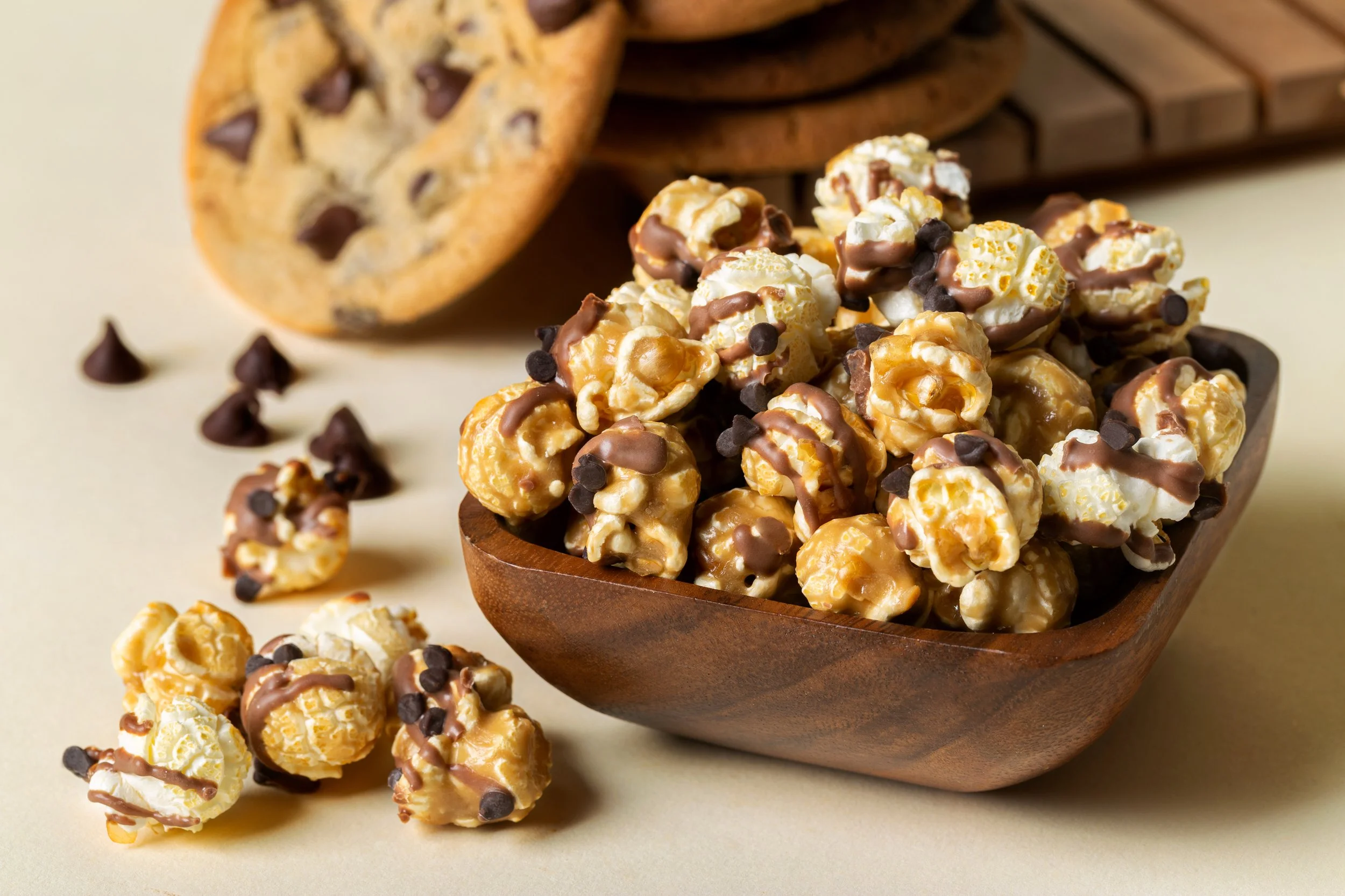 Caramel popcorn with chocolate drizzles in a wooden bowl, scattered on a beige background with a chocolate chip cookie in the background.