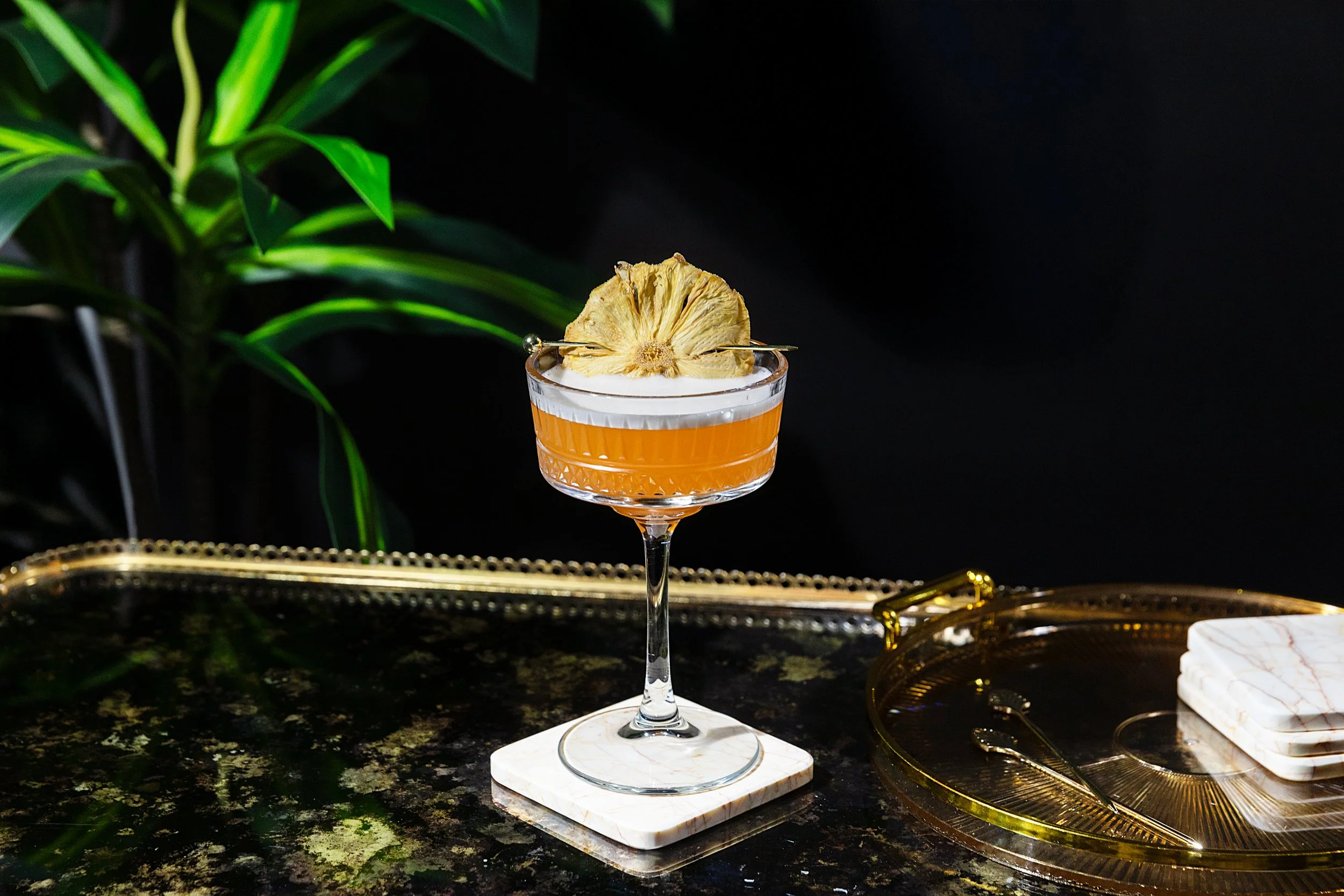 A cocktail glass filled with an orange-colored drink garnished with a dried pineapple slice, set on a white marble coaster on a dark, textured surface with green plants and gold decorative items in the background.