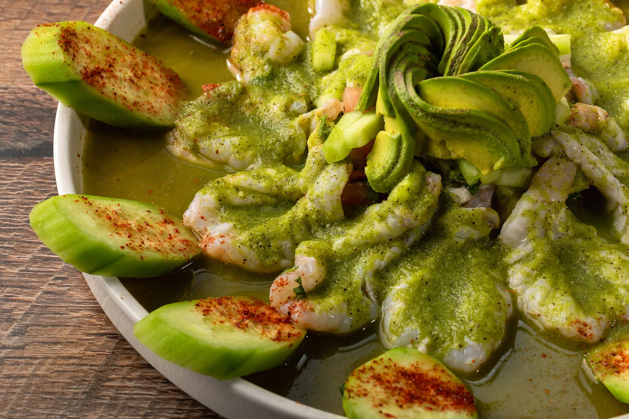 Close-up of a bowl of avocado and cucumber slices, topped with green sauce and sprinkled with red chili powder.