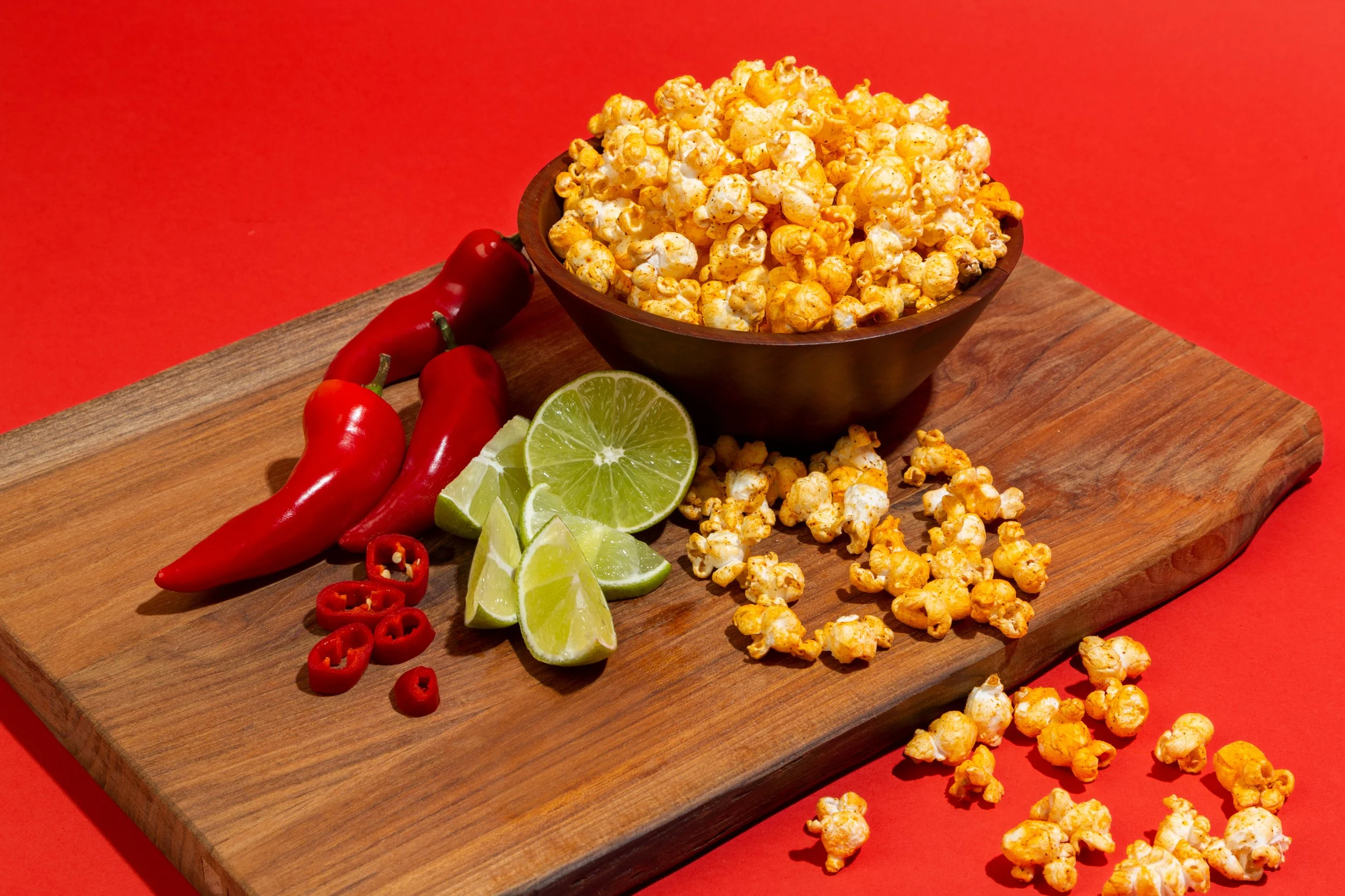 A wooden cutting board with two sliced lime wedges, three red chili peppers, and a bowl of buttered popcorn against a red background.