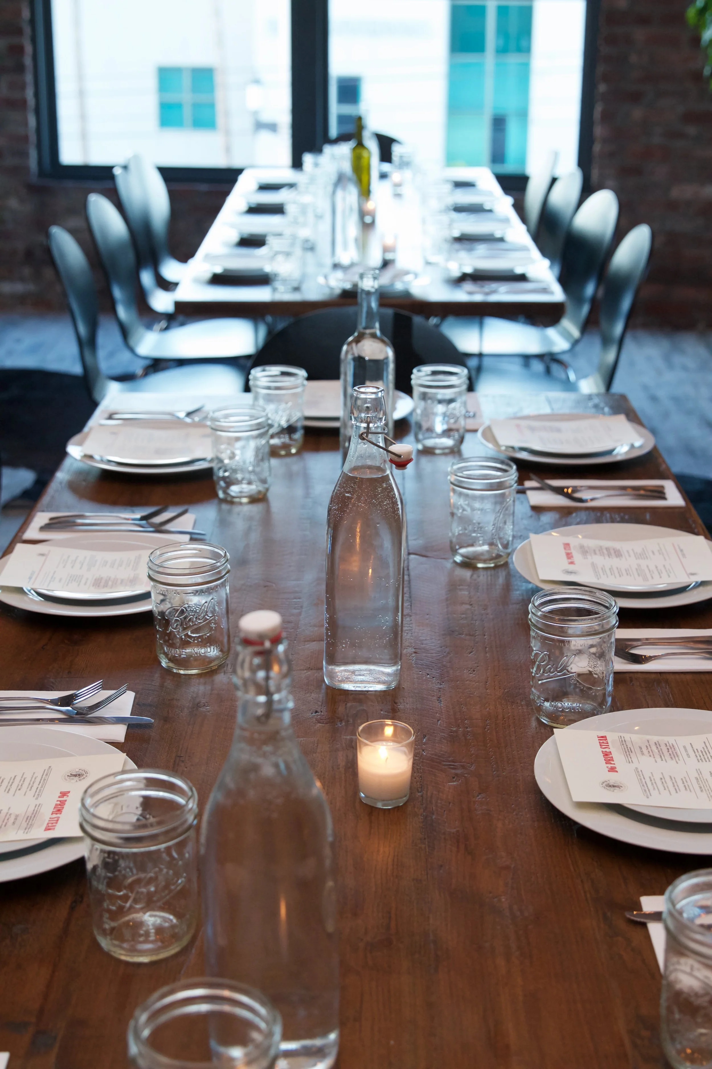 A long wooden dining table set for a meal in a bright, industrial-style room with large windows and exposed brick walls. The table includes plates, glasses, silverware, water bottles, and small candles.
