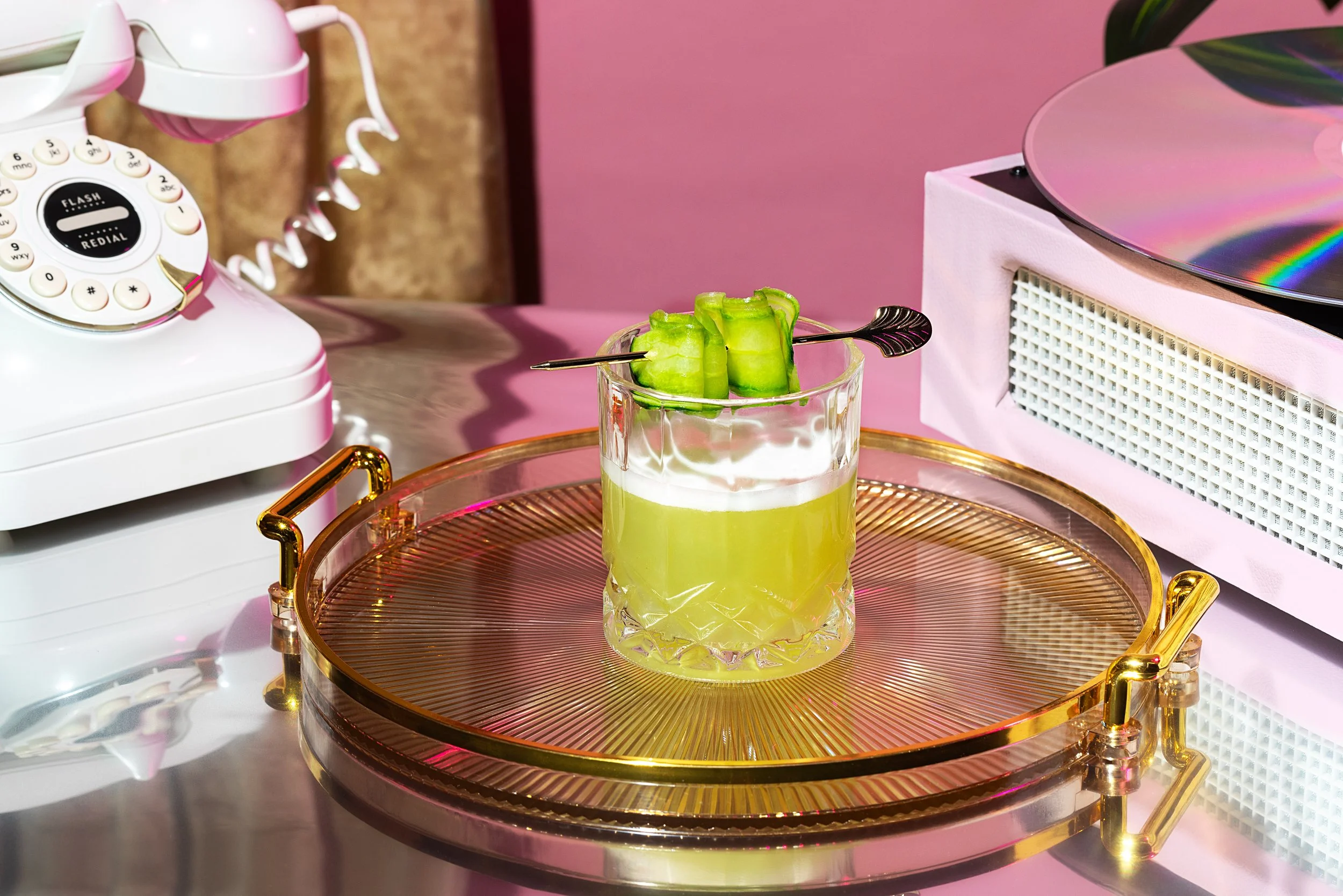 A glass filled with green juice garnished with cucumber slices and a cocktail pick is placed on a gold-accented tray. To the left, a vintage white rotary dial telephone is seen, and to the right, a turntable with a holographic vinyl record is partial