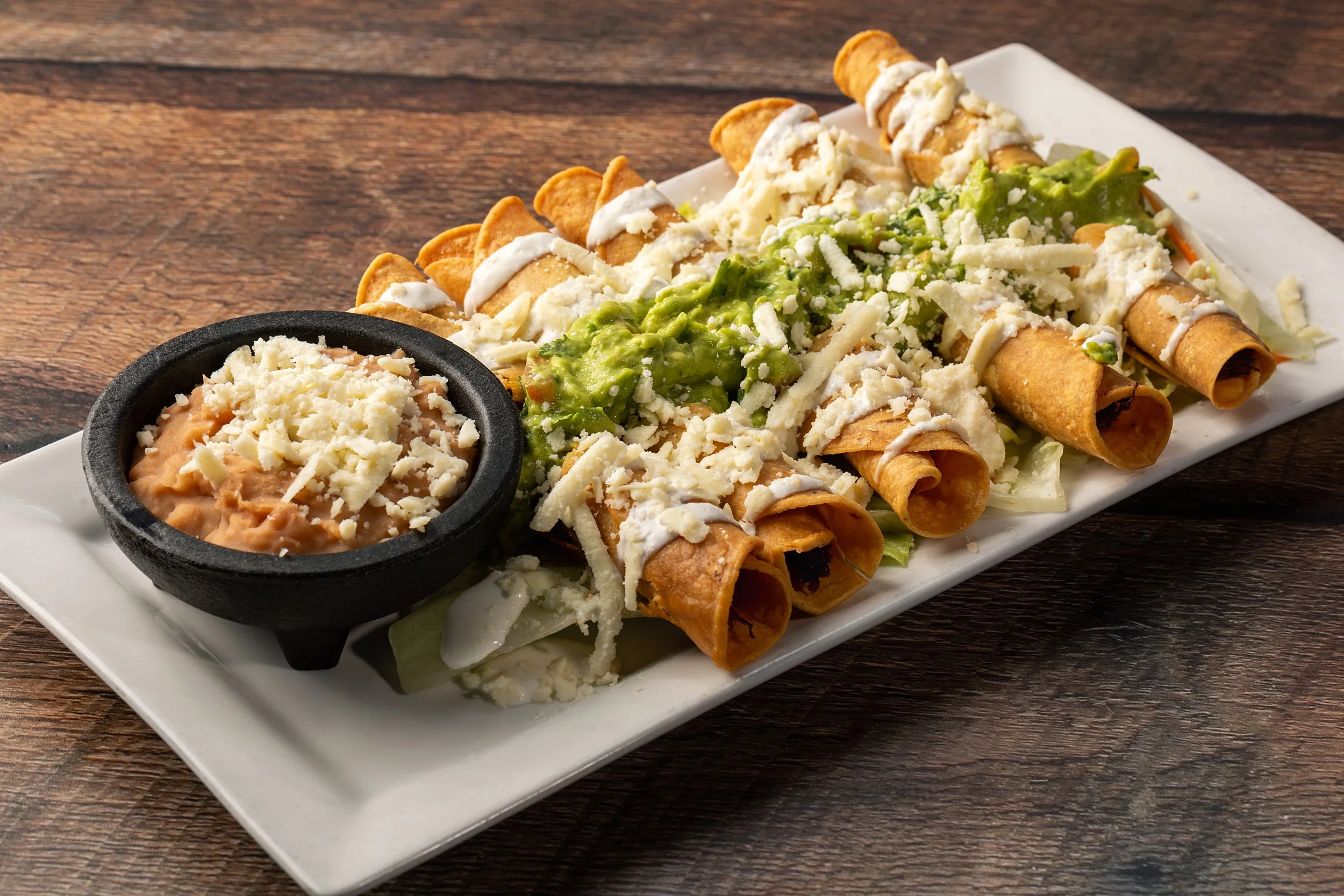 Plate of restaurant-style taquitos topped with shredded cheese, green guacamole, and drizzled with sour cream, served with a side of dipping sauce for tacos, on a white rectangular platter on a wooden table.