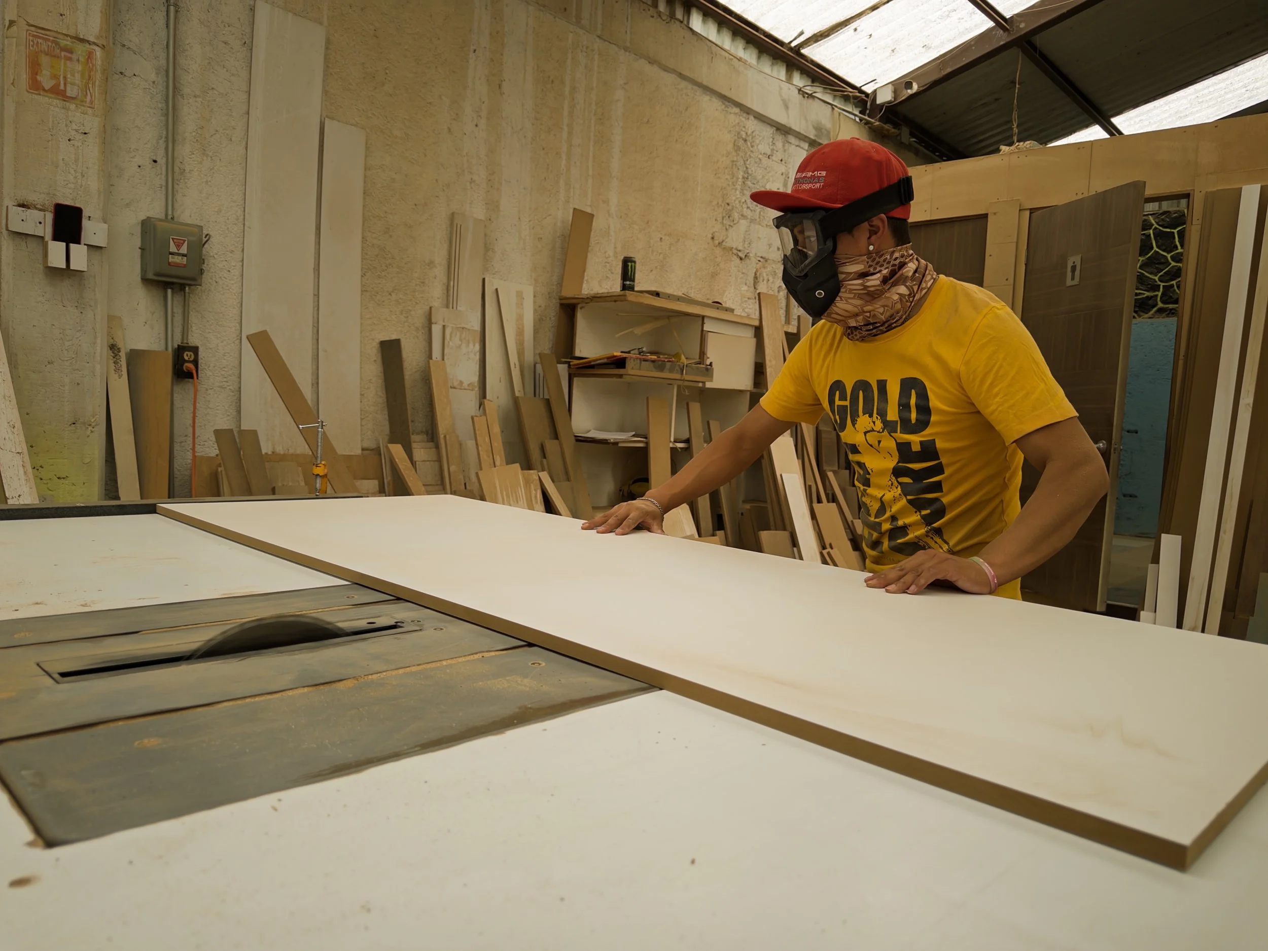 Joven trabajando en una carpintería con madera y herramientas, vistiendo gafas protectoras, gorra roja y camiseta amarilla en un taller de carpintería.