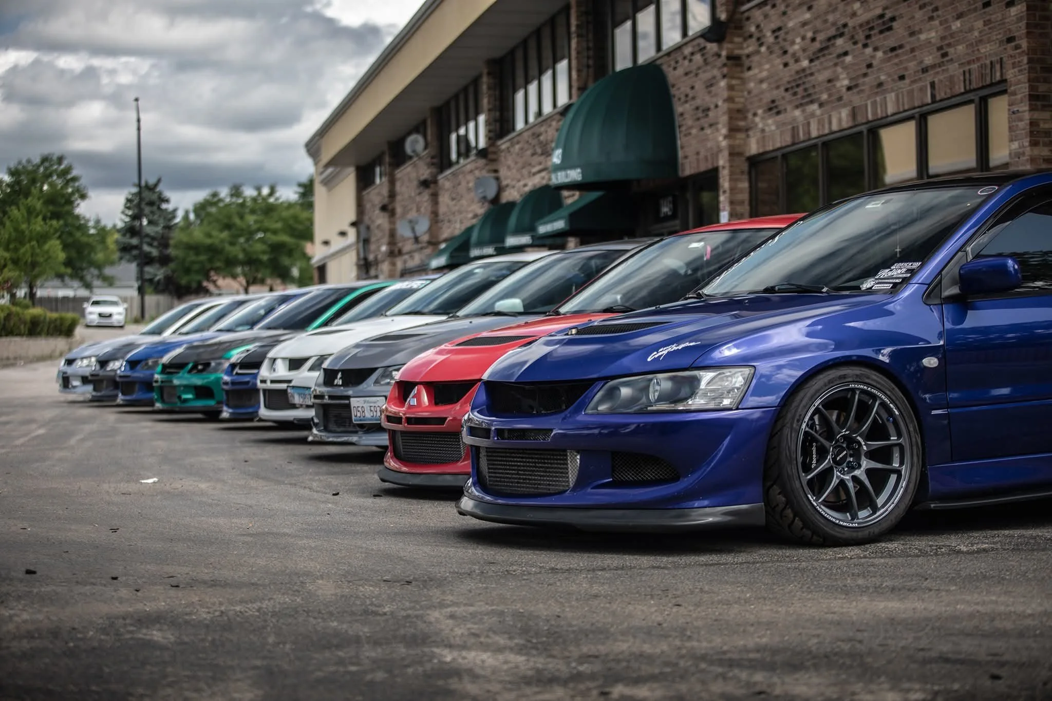 A row of colorful sports cars parked in front of a commercial building under a cloudy sky.