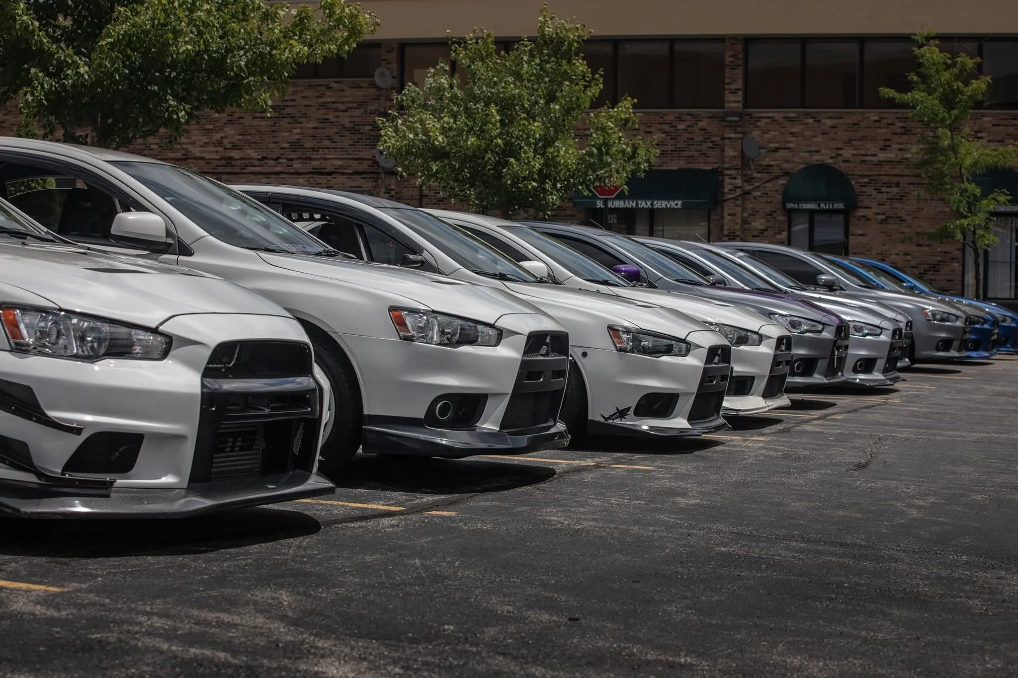A row of parked cars in a parking lot near a brick building with trees in front.
