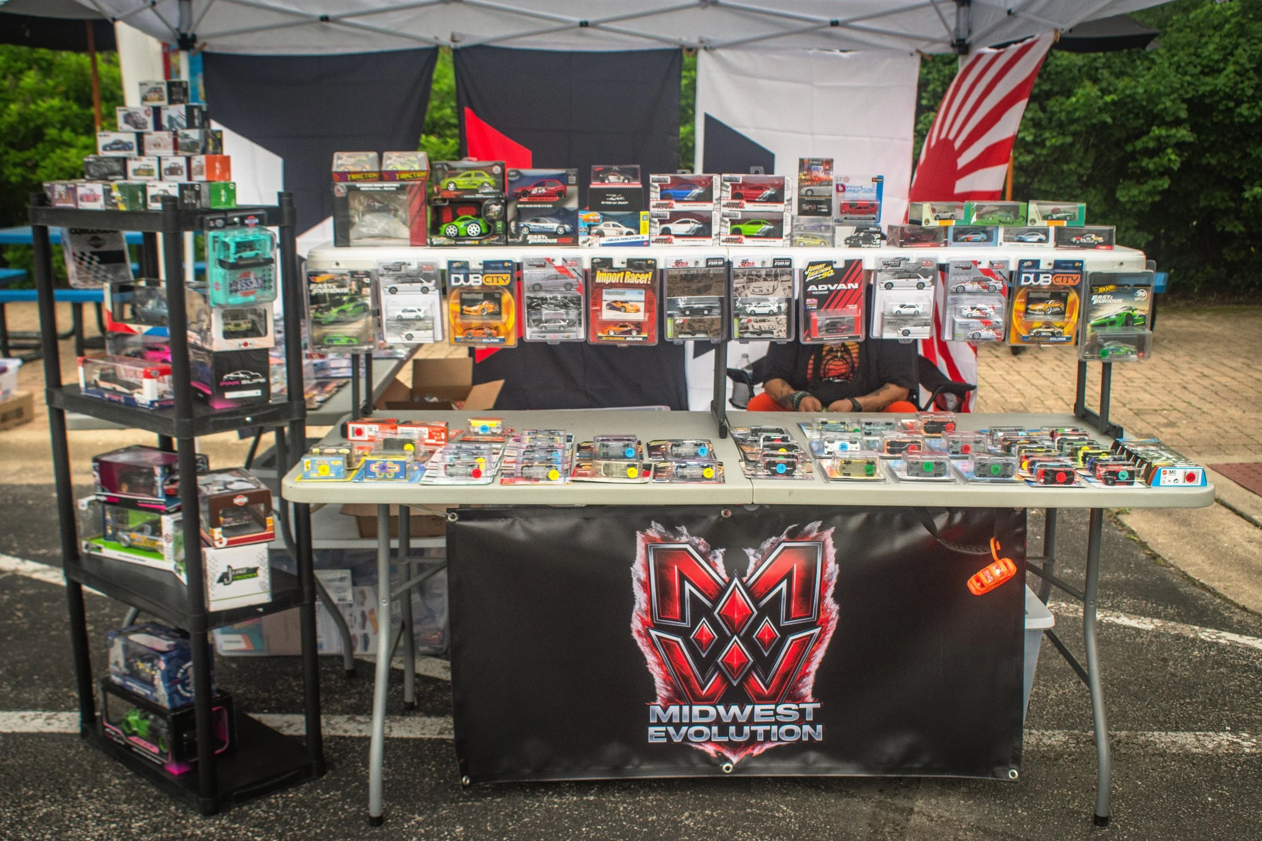 A table and shelves displaying collectible die-cast toy cars and models at an outdoor market stall with Midland Evolution branding and a backdrop of flags and greenery.