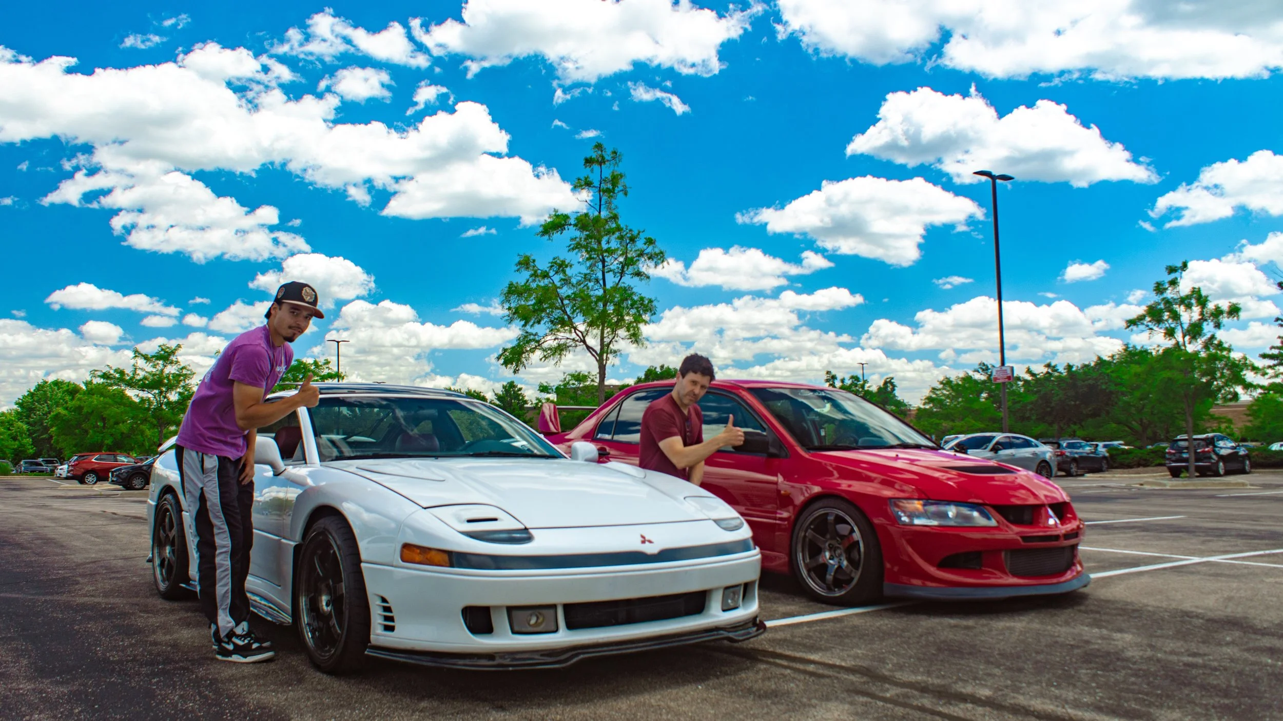 Two men standing next to sports cars in a parking lot under a partly cloudy sky. One man in a purple shirt and cap giving a thumbs up next to a white car, and the other in a red shirt giving a thumbs up next to a red car.
