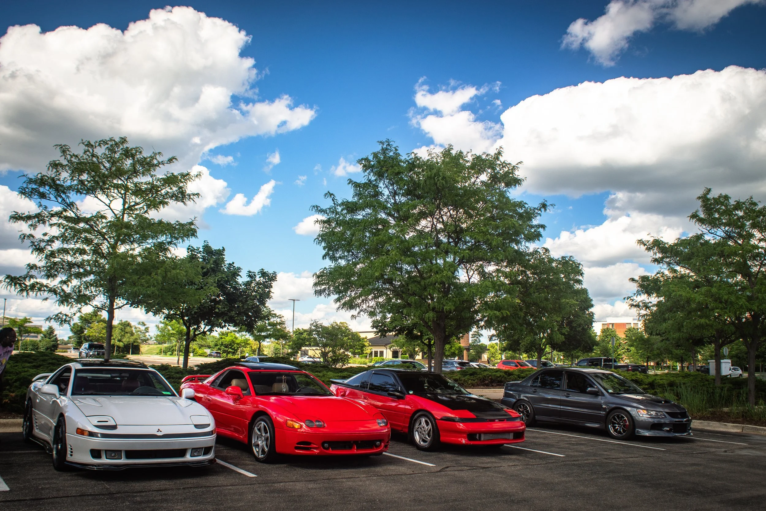 A parking lot filled with sports cars, with trees and a blue sky with white clouds in the background.