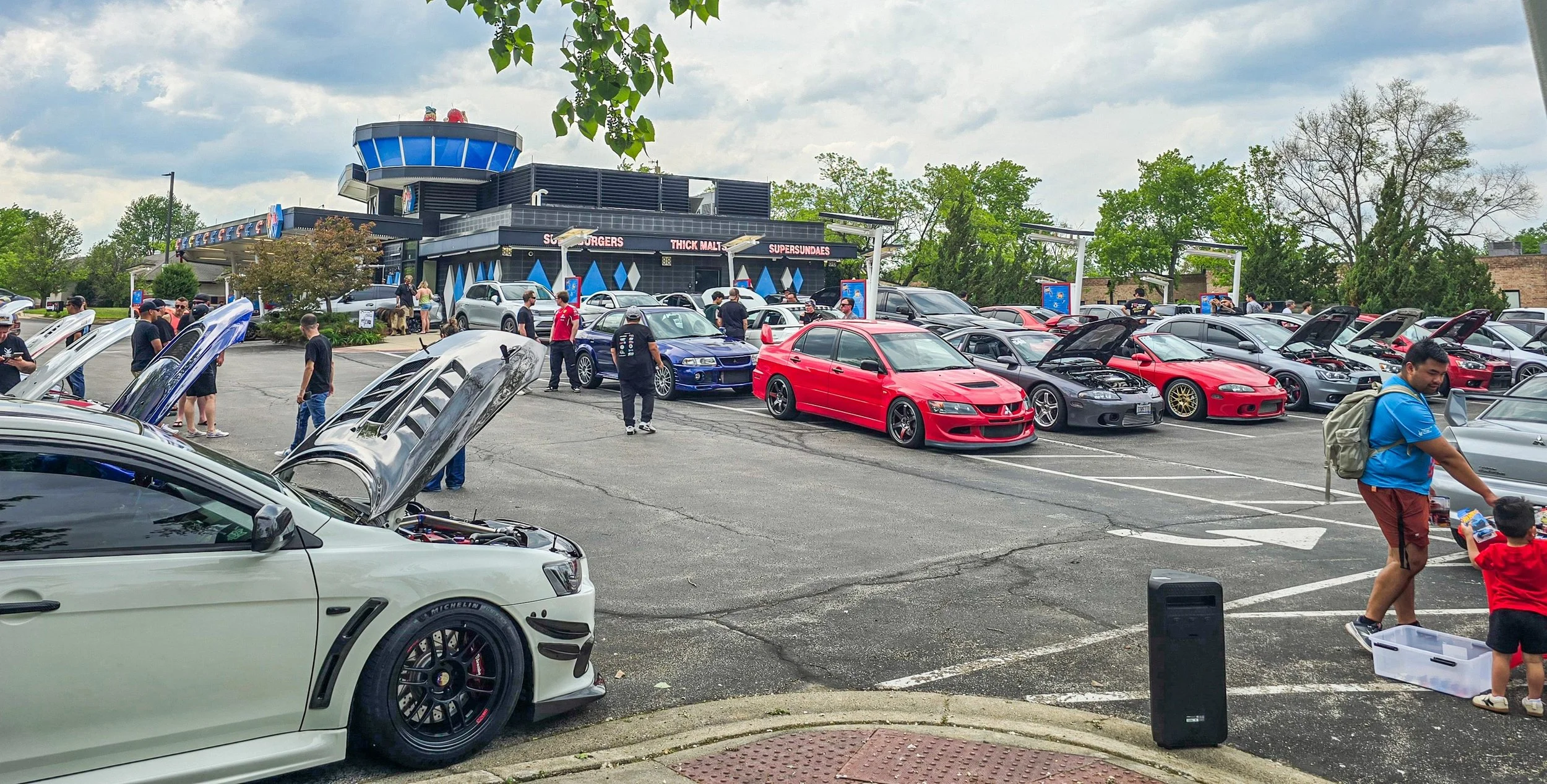 Car meet in a parking lot with various sports cars, some with hoods open, and attendees walking around and socializing, near a fast food restaurant.