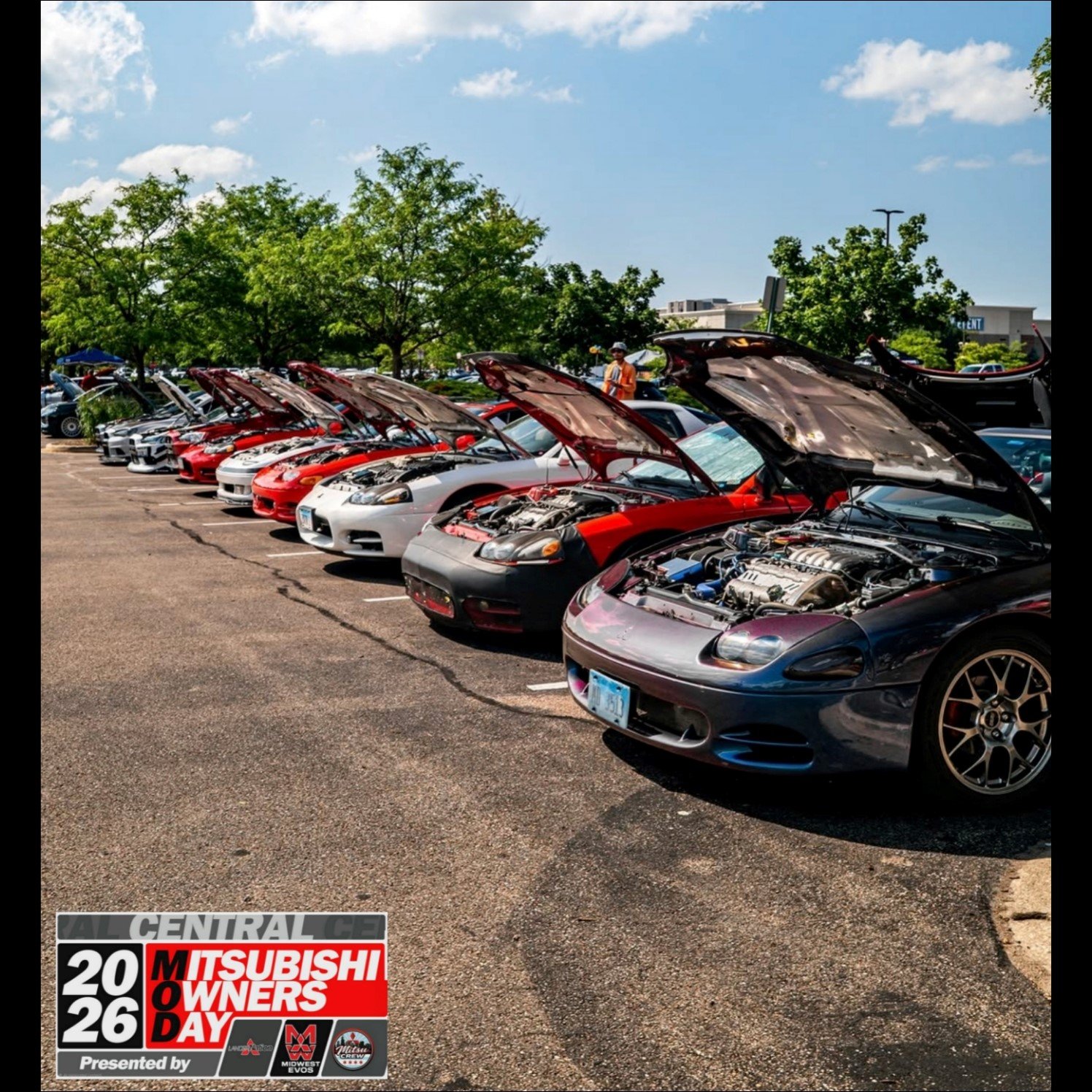 A line of sports cars with their hoods open at a car show on a sunny day, with trees and buildings in the background.