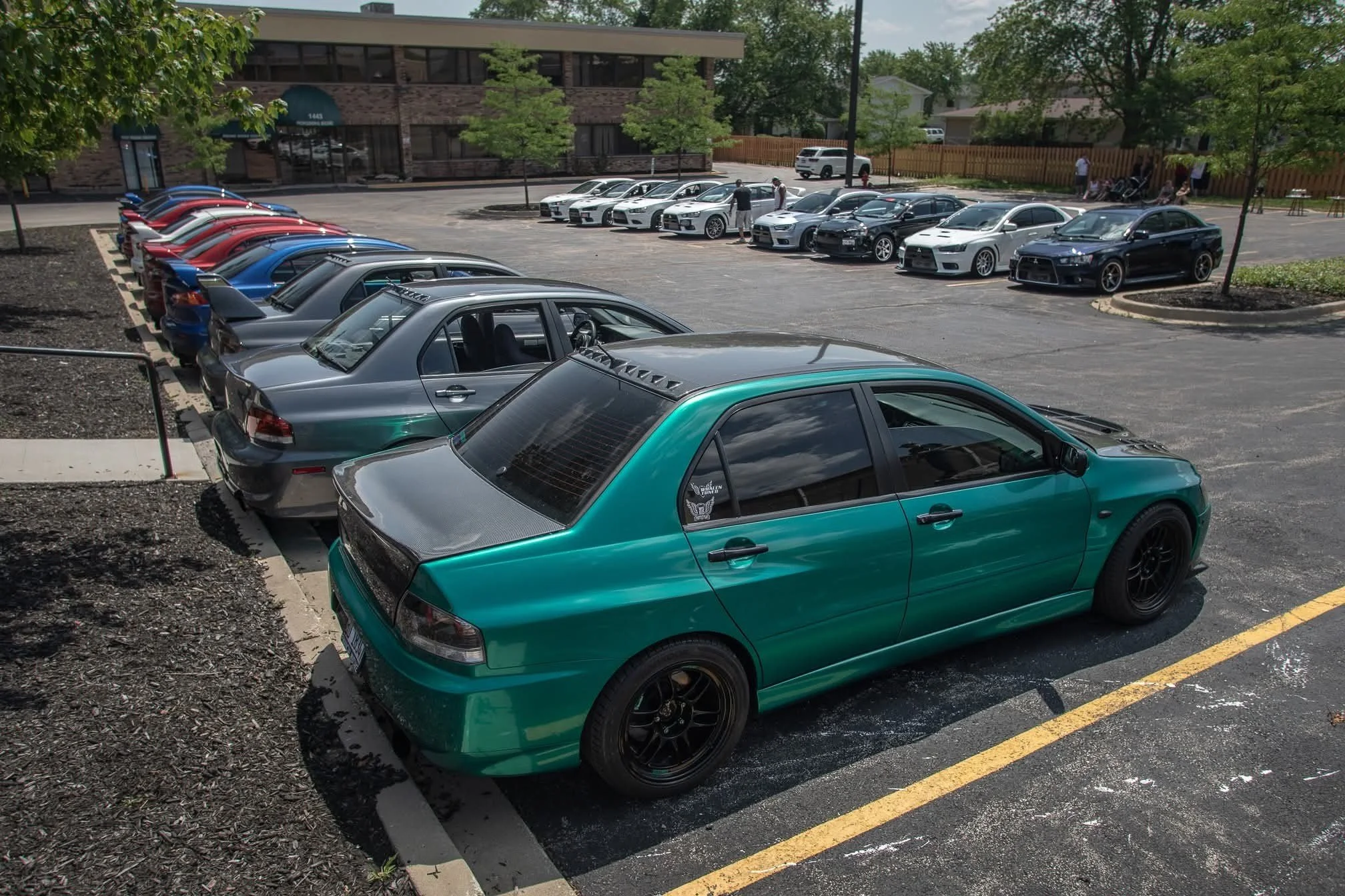 A parking lot with various parked cars, including a green car in the foreground and a lineup of sports cars and sedans in the background. There are a few people walking and standing near the cars, and trees and a building are visible in the surroundings.