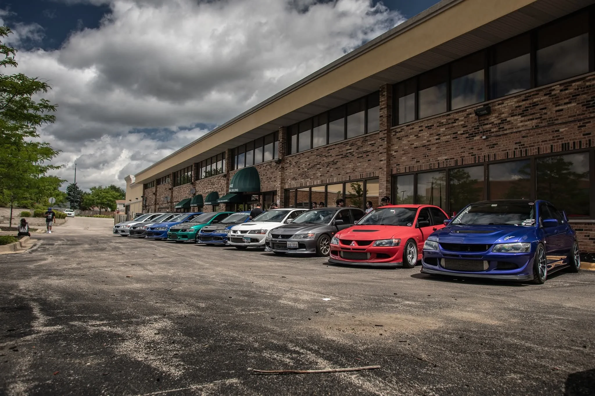 A parking lot with sports cars lined up in front of a brick building under a cloudy sky.