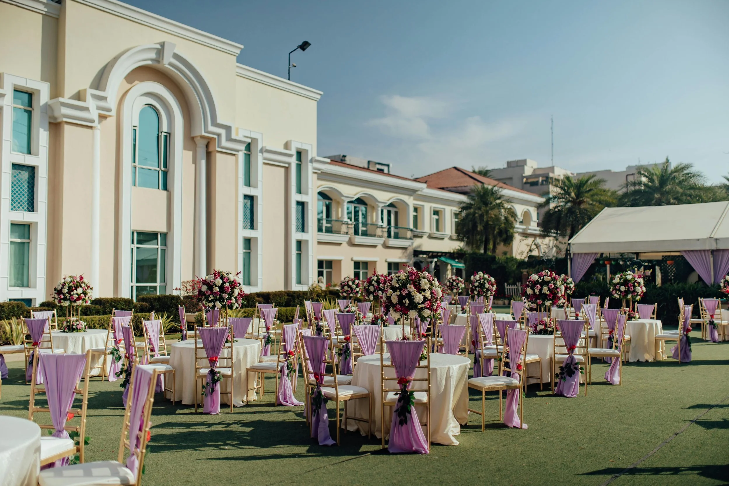 Outdoor wedding reception setup with round tables covered in white tablecloths, decorated with purple chair sashes and floral centerpieces. The setting is in front of a cream-colored building with arched windows and balconies, with a blue sky overhead and palm trees in the background.