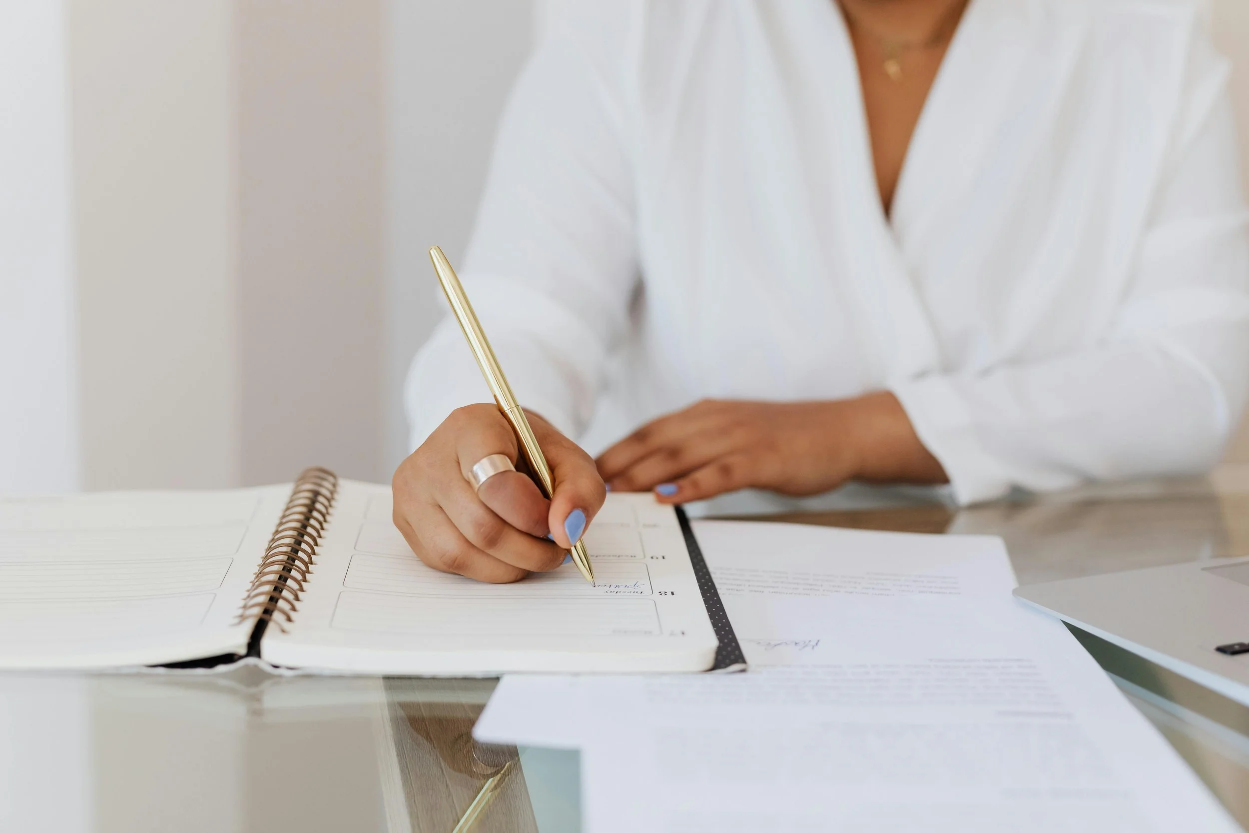 A person writing in a planner with a gold pen, sitting at a desk with papers and books.