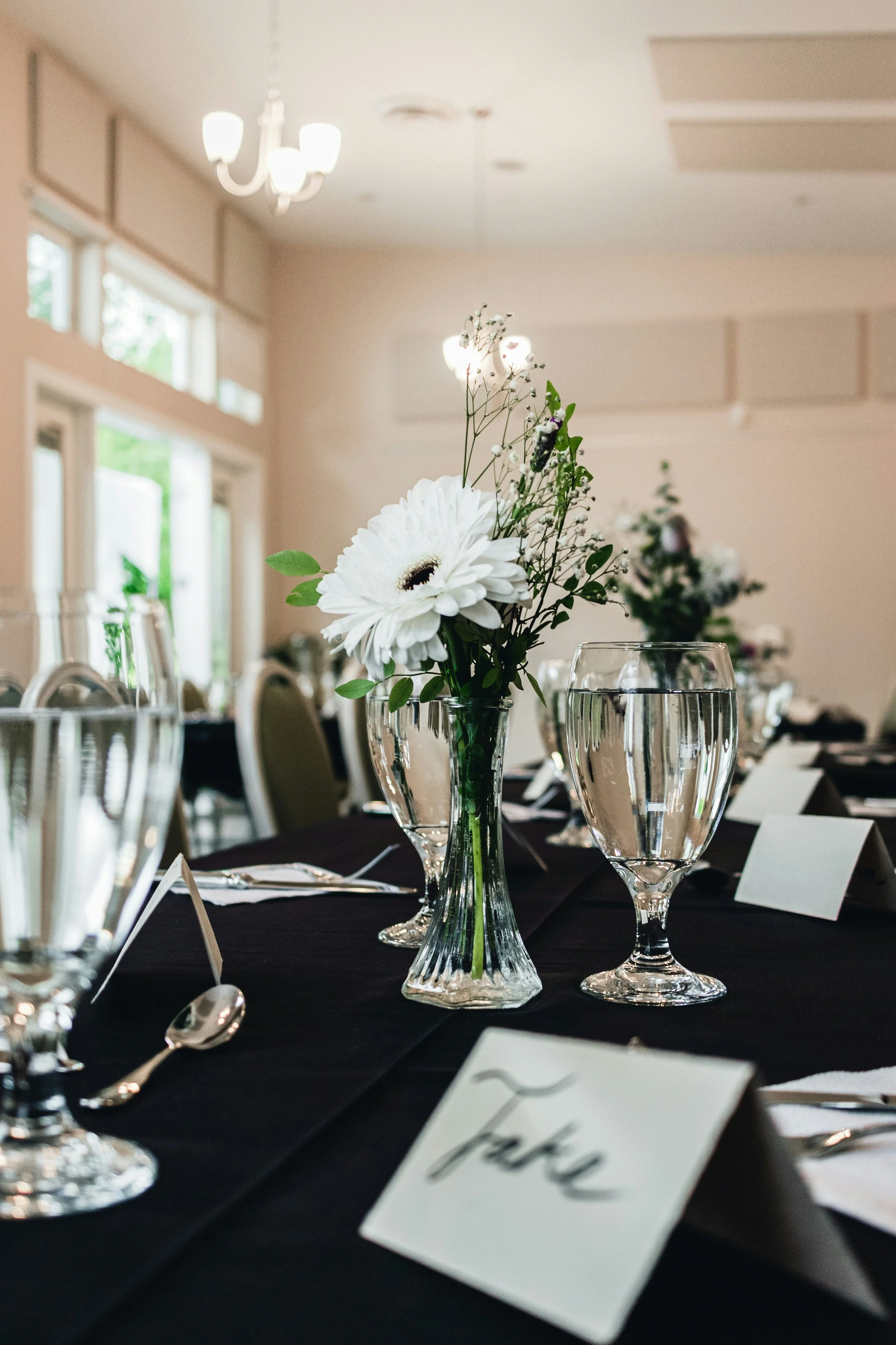 A table set with a black tablecloth, glassstemmed wine glasses, silverware, place cards, and a centerpiece with white daisy-like flowers and greenery, in a well-lit room with large windows and modern ceiling lights.
