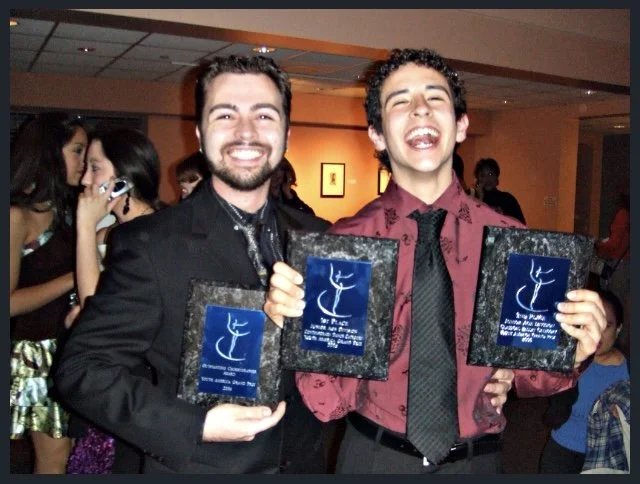 Two men smiling and holding awards at an event, with other people in the background.
