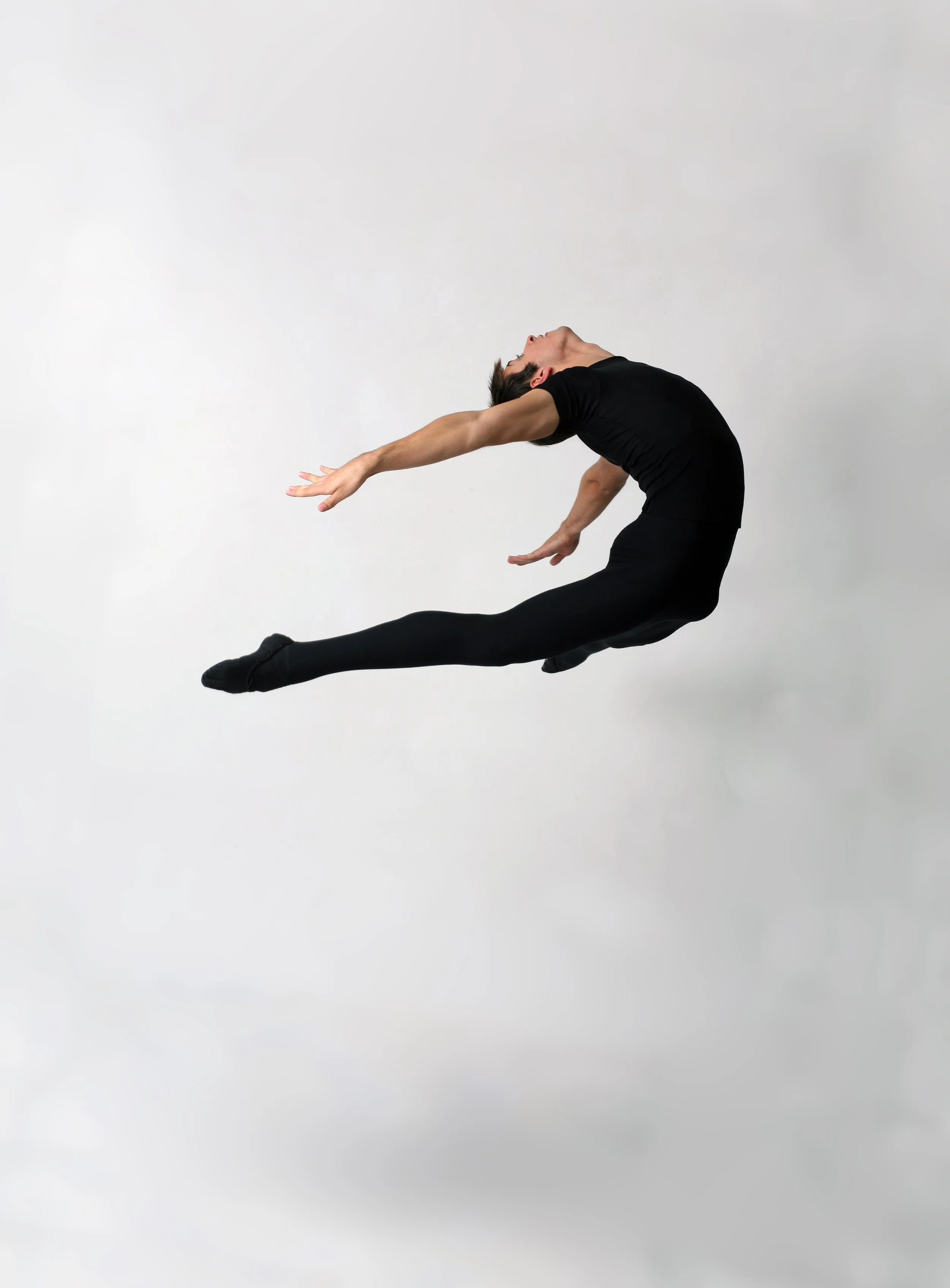 A woman performing a dance pose with her back arched, arms extended, and one leg straight out, dressed in black against a plain white background.