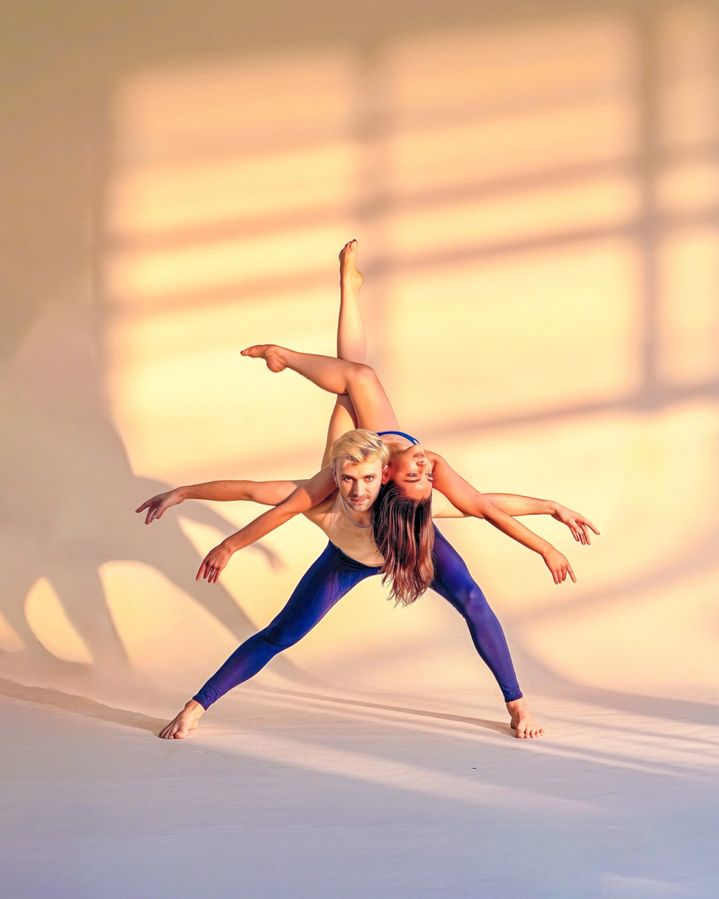 A male and female dancer perform a balancing act together, with the woman's legs extending upward and the man's face close to the floor, against a warm, gradient background with shadows.