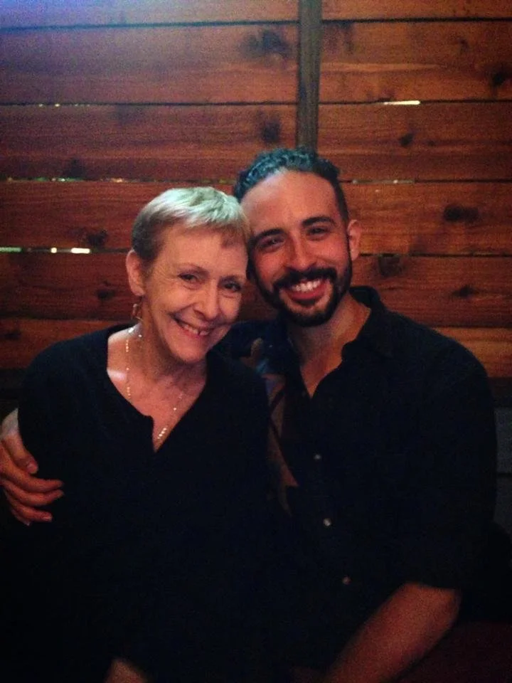 A smiling older woman and a smiling younger man sitting close together in a wooden room, possibly a restaurant or cabin, sharing a happy moment.