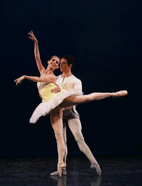 Two ballet dancers perform on stage, with one supporting the other, who is executing a high leg extension. The ballerina wears a yellow tutu and pointe shoes, while the male dancer wears white tights and a white shirt. The background is dark.