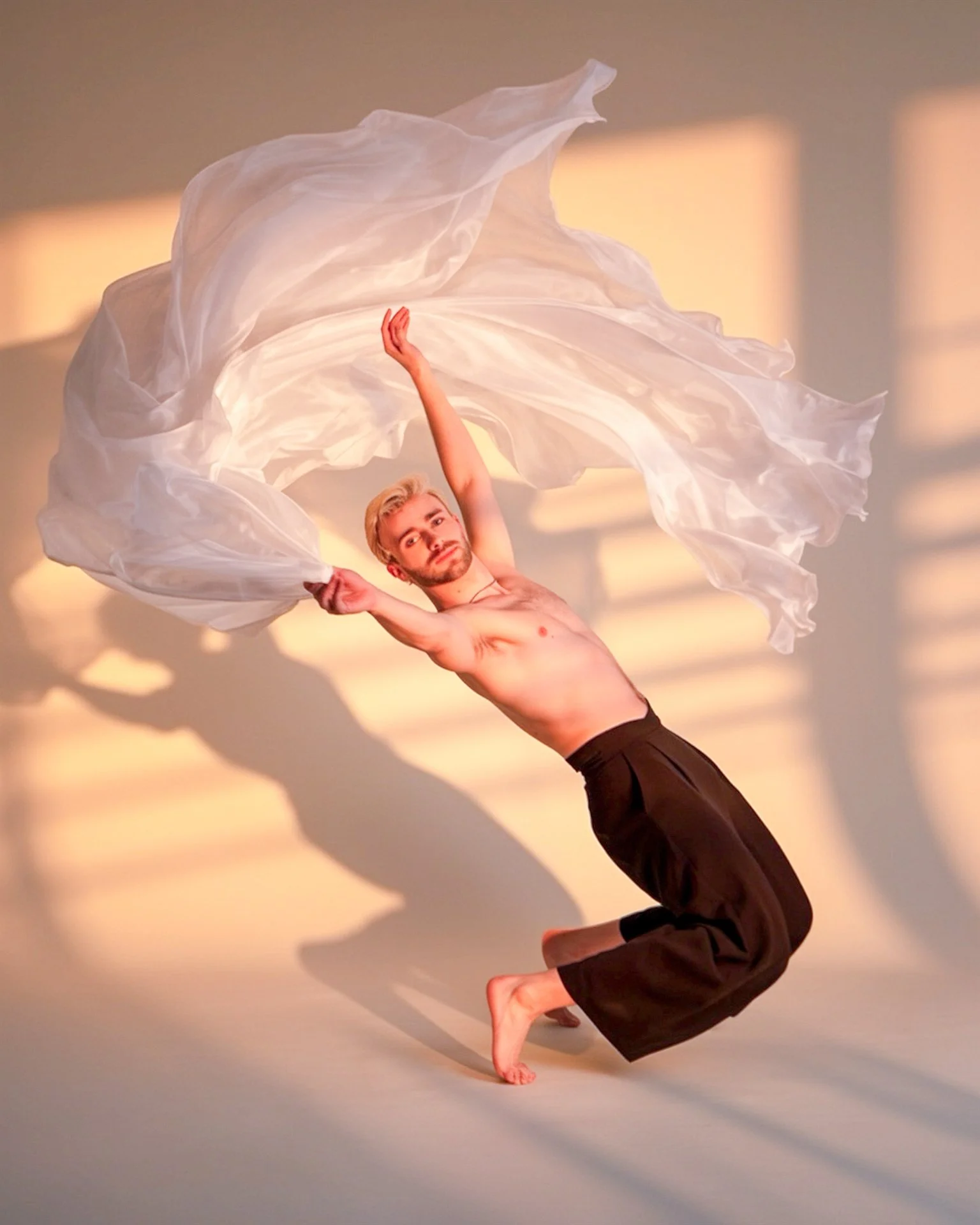 A man performs a handstand with his body bent at the hips and his legs elevated, spinning a large, flowing piece of fabric in the air. The background has warm, soft lighting and shadows.