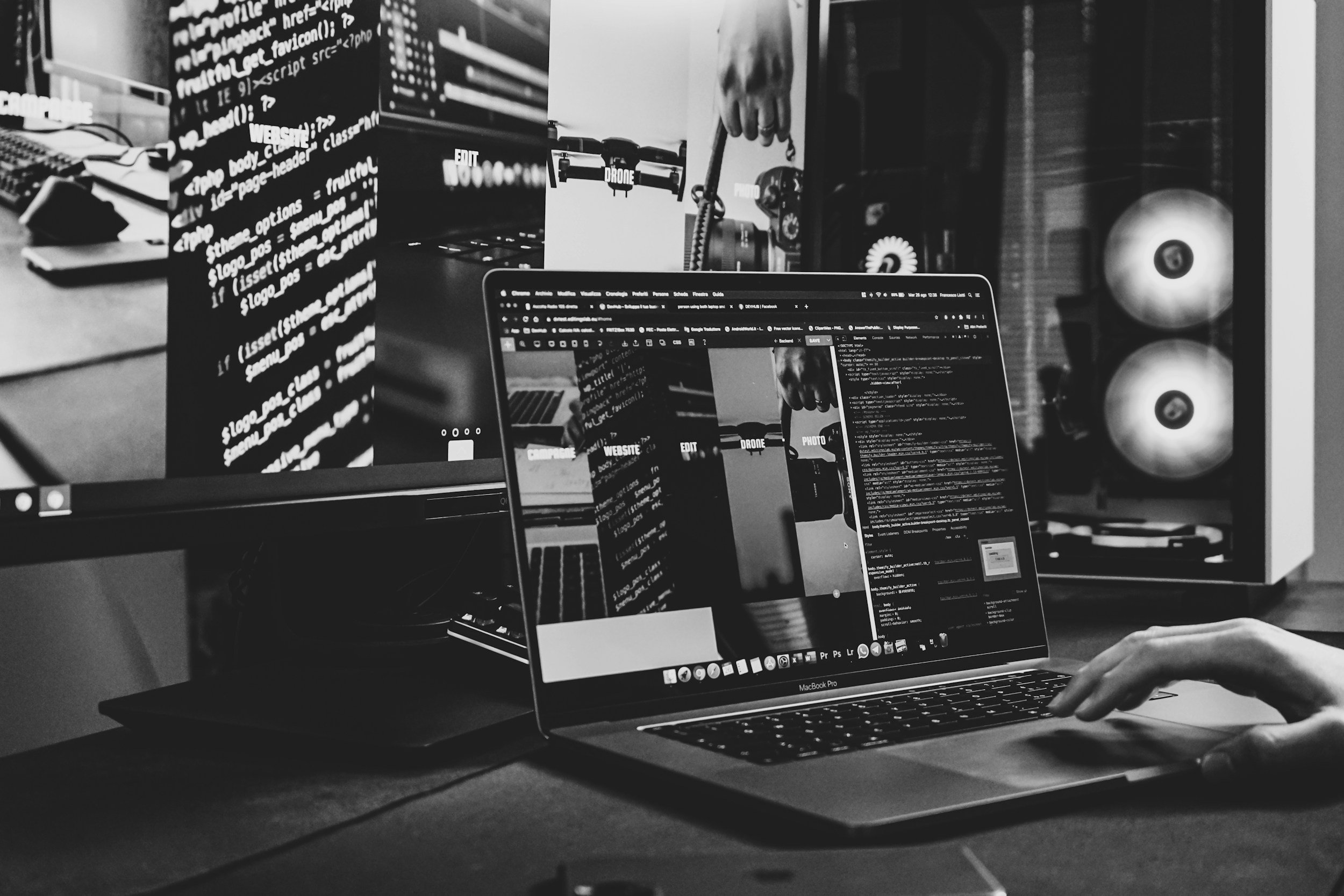 Black and white photograph of a person working on a laptop in an office or tech workspace. The screen displays coding or web development. Multiple monitors and technical equipment, including a drone, are visible in the background.