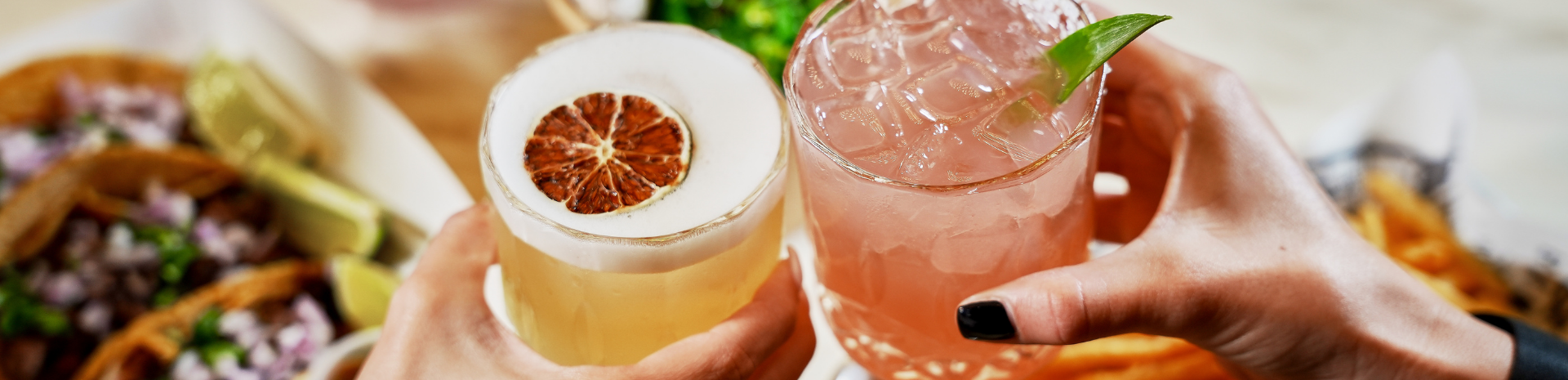 Two hands holding glasses of drinks, one with a lemon-lime soda and a dried lemon slice, the other with a pink cocktail garnished with a lime wedge, blurred food in the background.