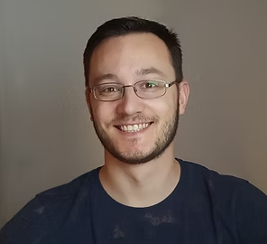 A smiling man with glasses and a beard wearing a navy blue shirt, standing against a plain gray background.