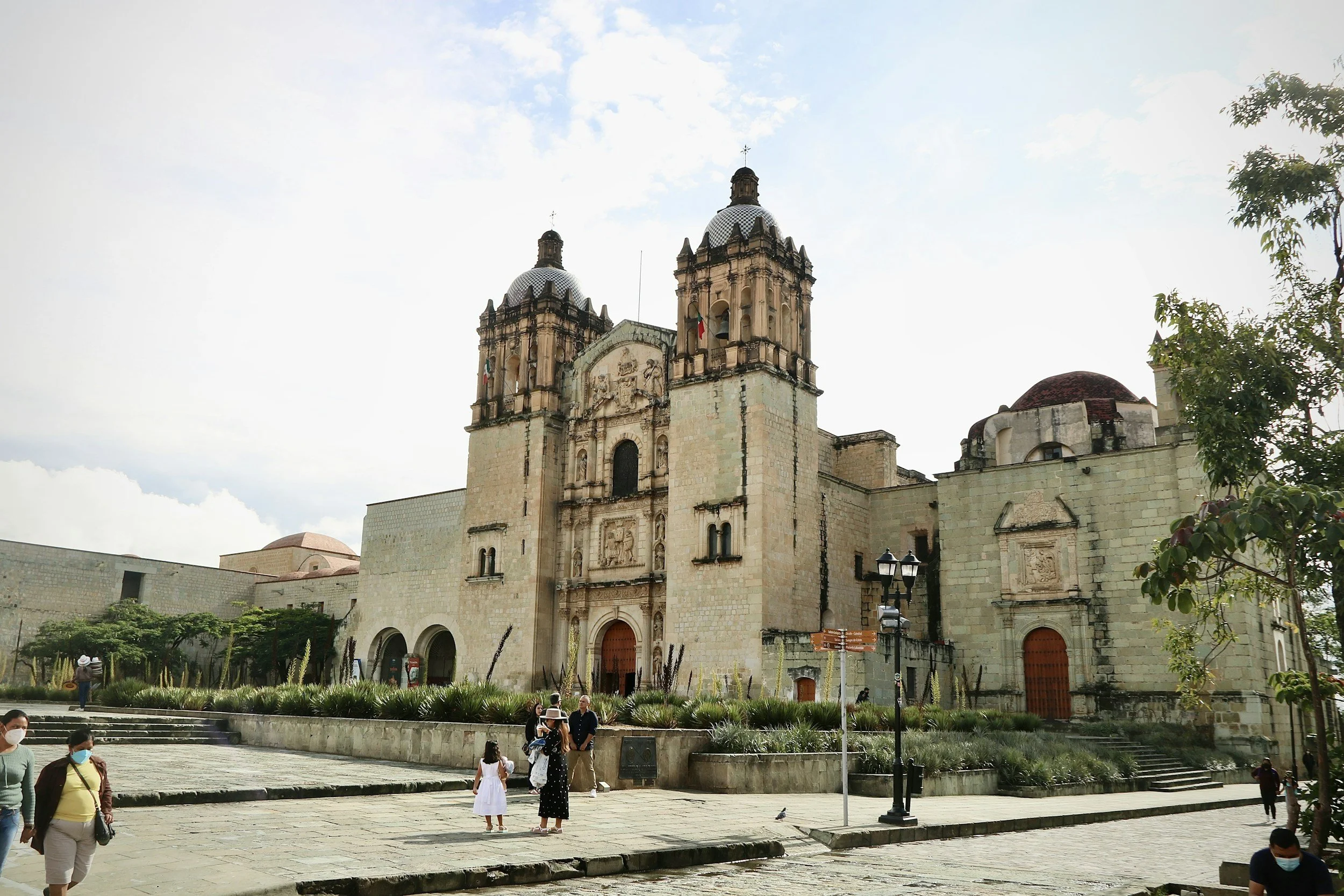 Vista de una iglesia colonial de piedra con dos torres y un frontón decorado en el centro, en una plaza con personas y árboles alrededor.