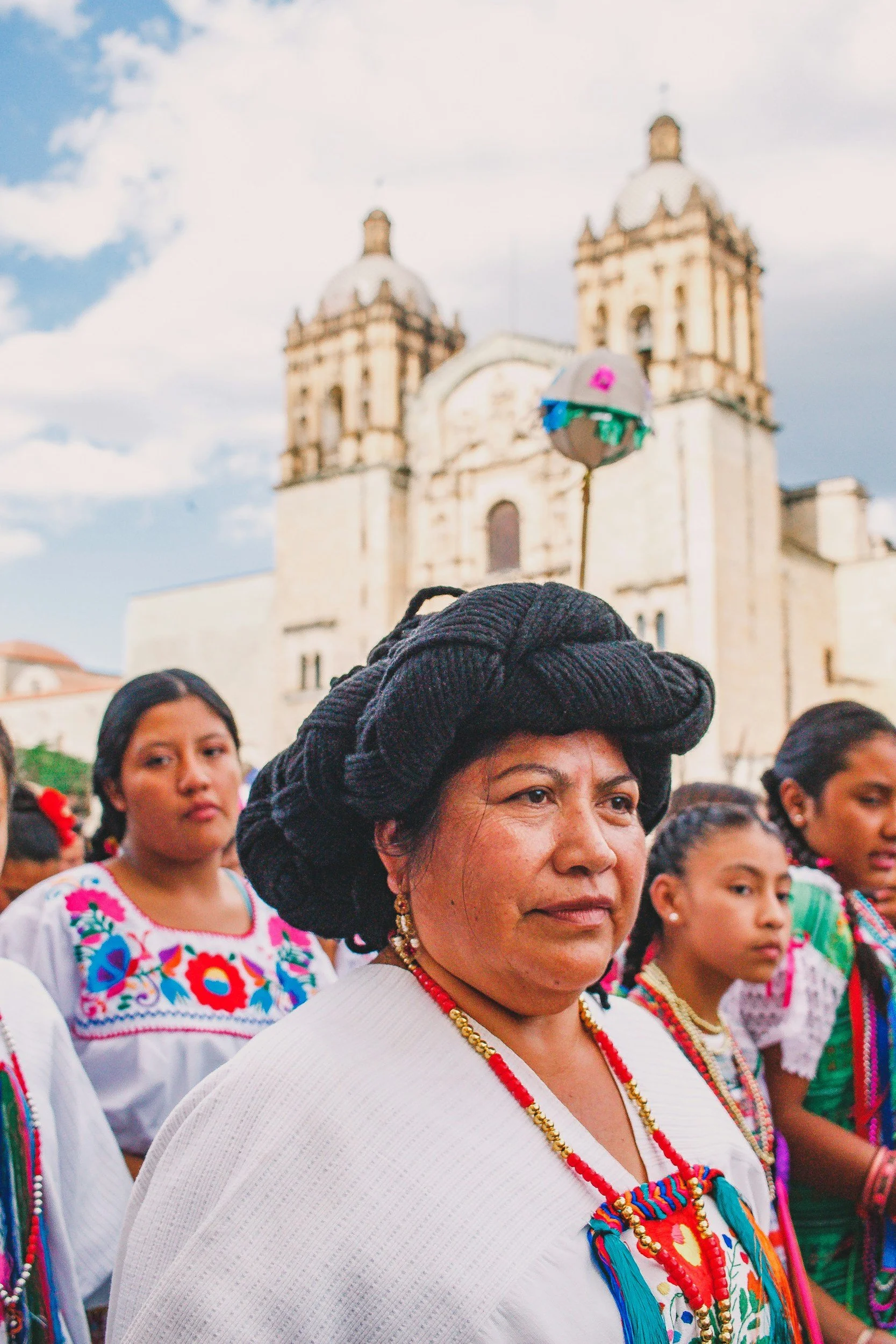 Grupo de mujeres indígenas con trajes tradicionales en una celebración en un lugar con una iglesia histórica de fondo bajo un cielo nublado.