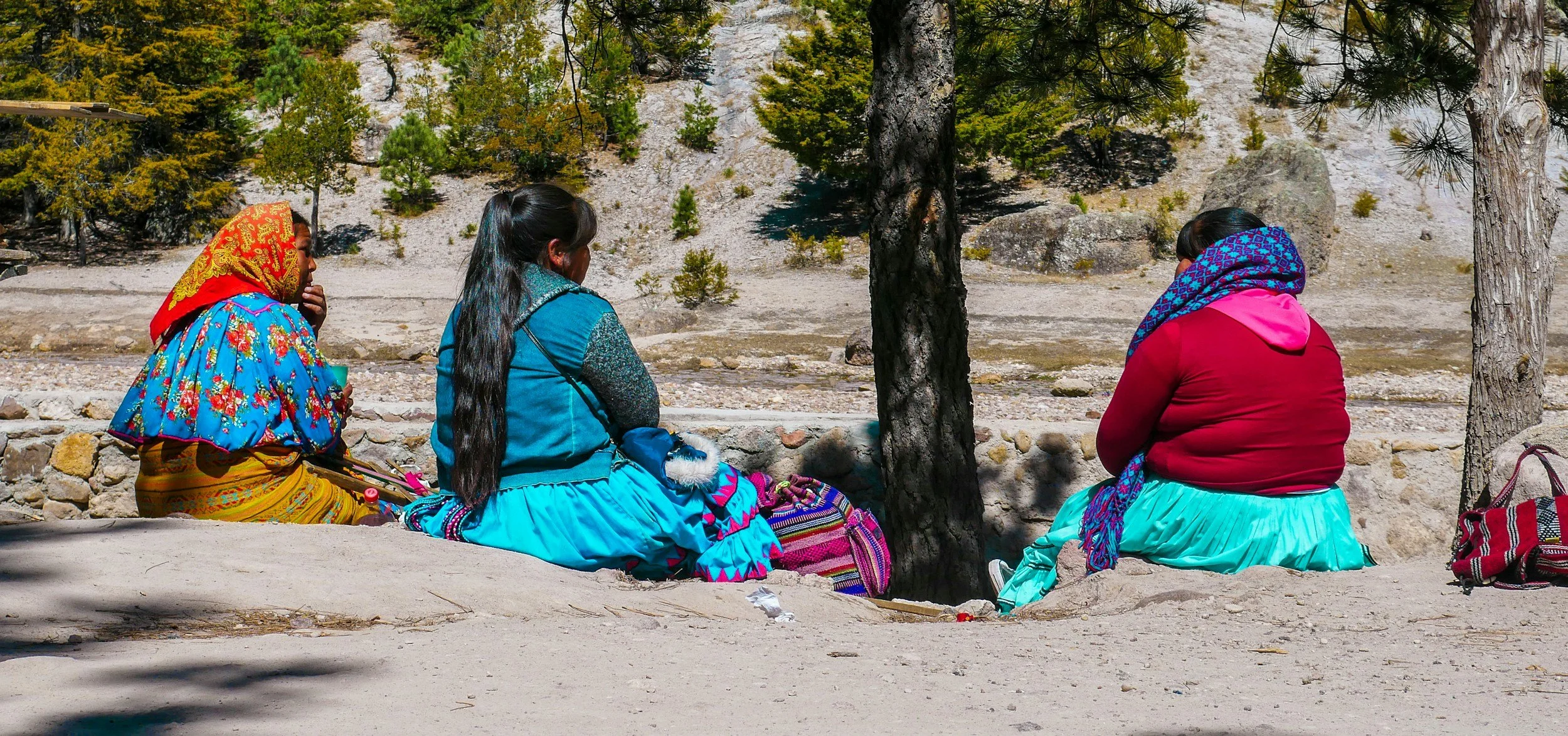 Tres mujeres indígenas sentadas en un muro de piedra en un ambiente natural, rodeadas de árboles y rocas, vestidas con ropa tradicional colorida y coloridas mantas.