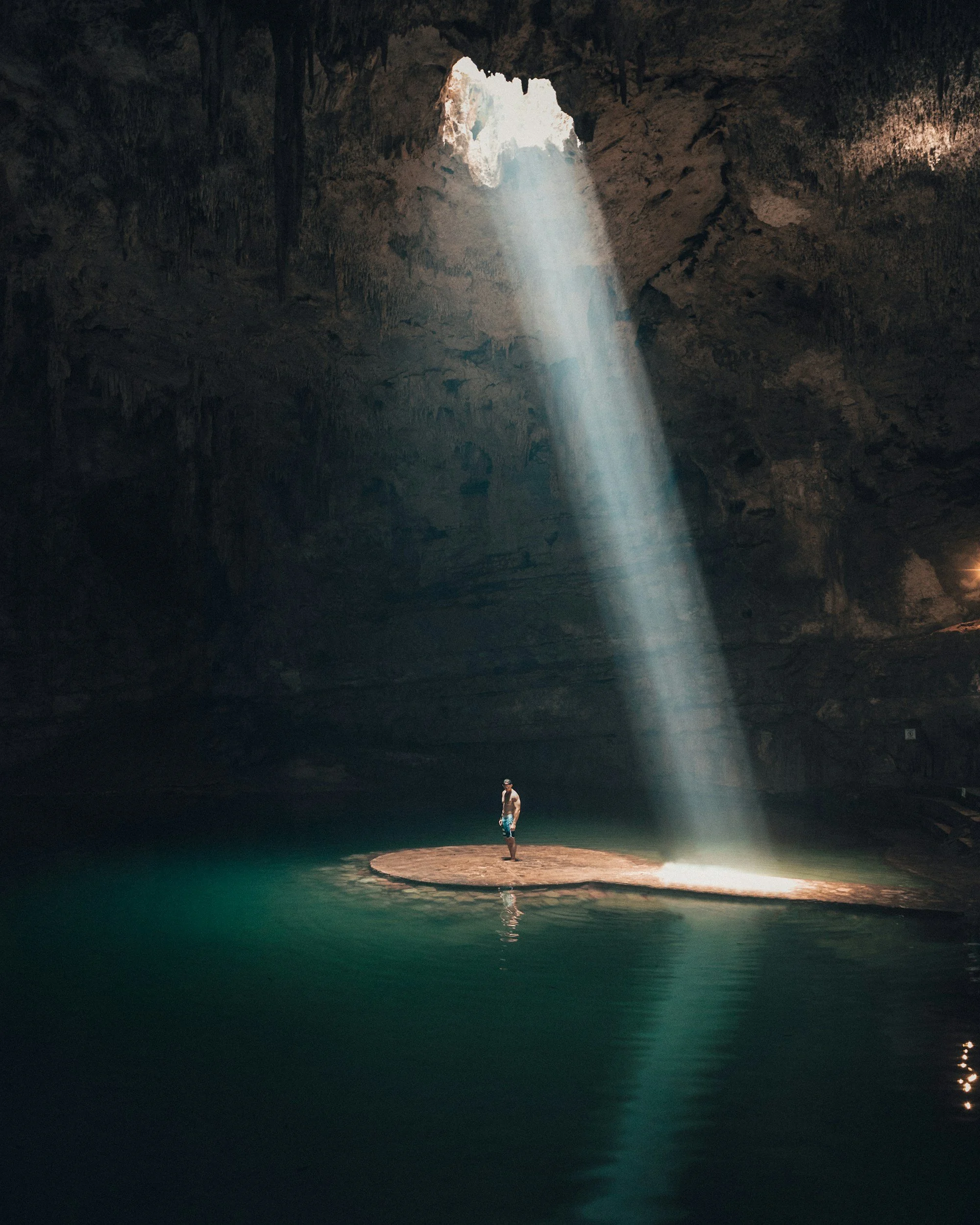 Una persona de pie sobre una roca en un lago dentro de una cueva, con un rayo de luz que entra por un agujero en el techo, iluminando a la persona y el agua circundante.