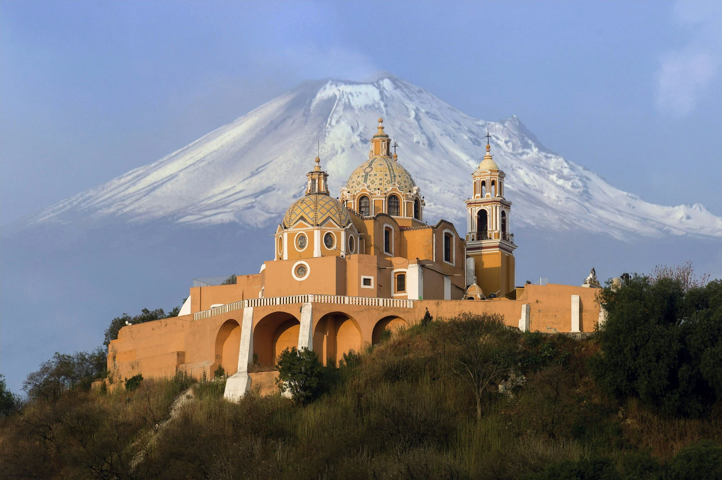 Vista panorámica de una iglesia colonial color naranja con cúpulas y campanario, en primer plano, con el volcán Tacaná nevado en el fondo, bajo cielo despejado.