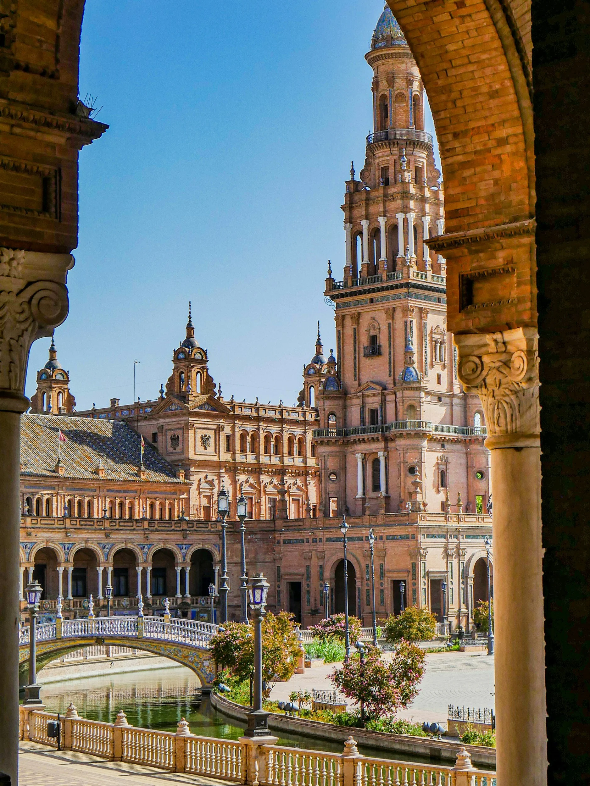 Vista de un edificio histórico y una torre en un clima soleado visto desde un balcón con columnas decoradas, un puente peatonal decorado y un río artificial en primer plano, rodeado de árboles y farolas.