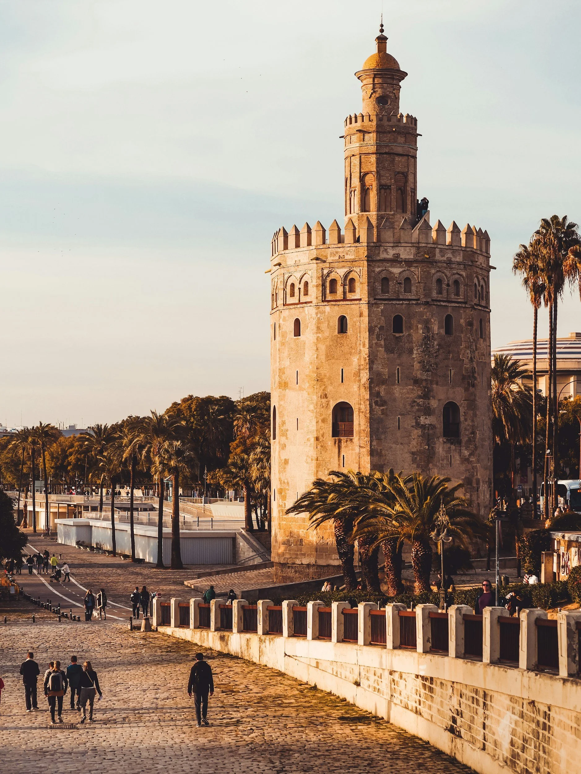 Torre histórica de piedra en el centro, rodeada de palmeras y personas caminando en un día soleado.