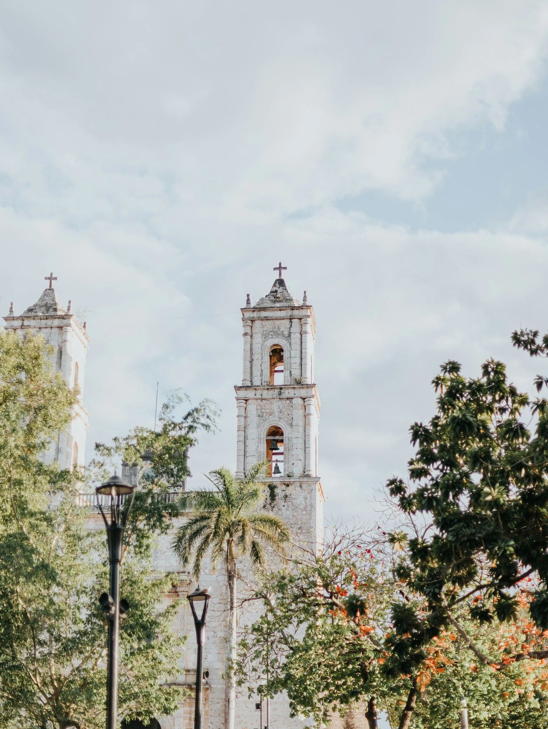 Torre de iglesia antigua con campanas y cruces en la parte superior, rodeada de árboles y faroles en un día nublado.