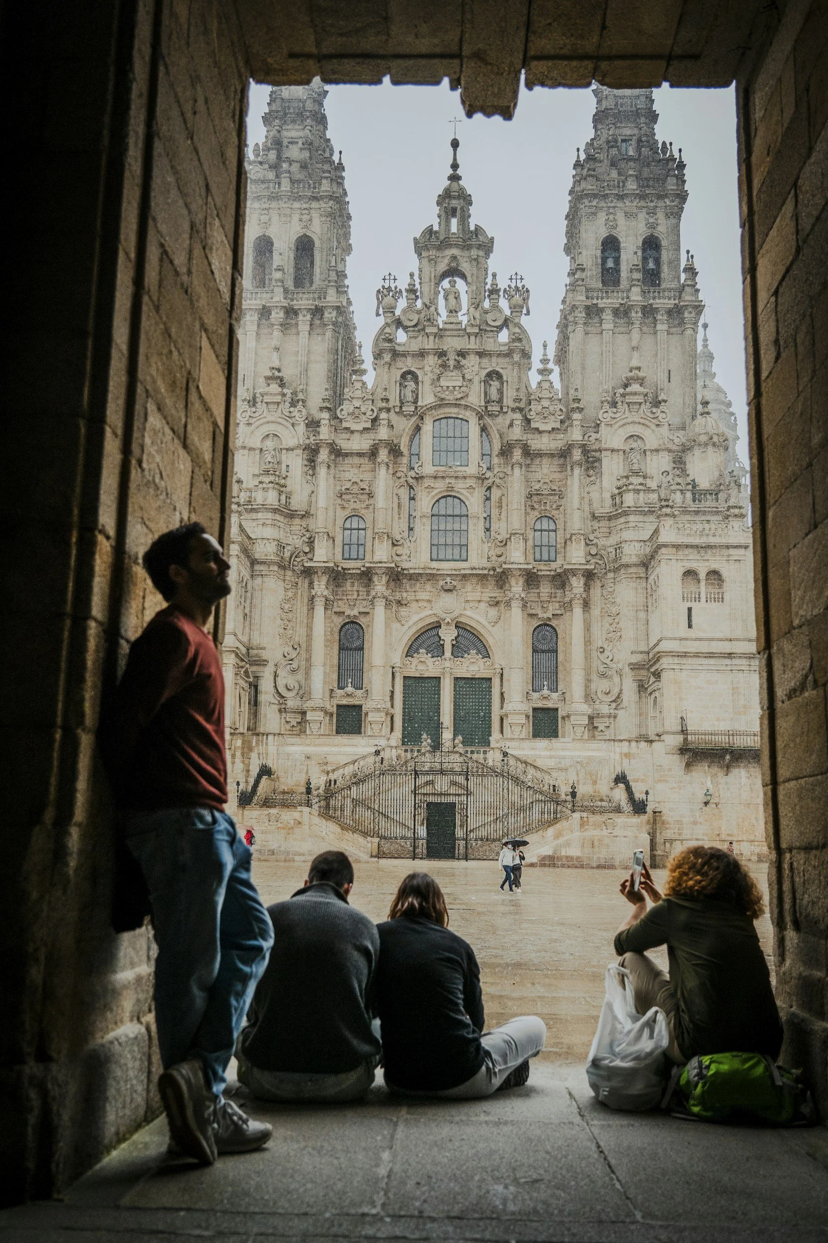 Vista de un colegio religioso con torres altas y detalles arquitectónicos elaborados, vista desde un pasillo con personas sentadas en el suelo y otras tomando fotos, en un día lluvioso.