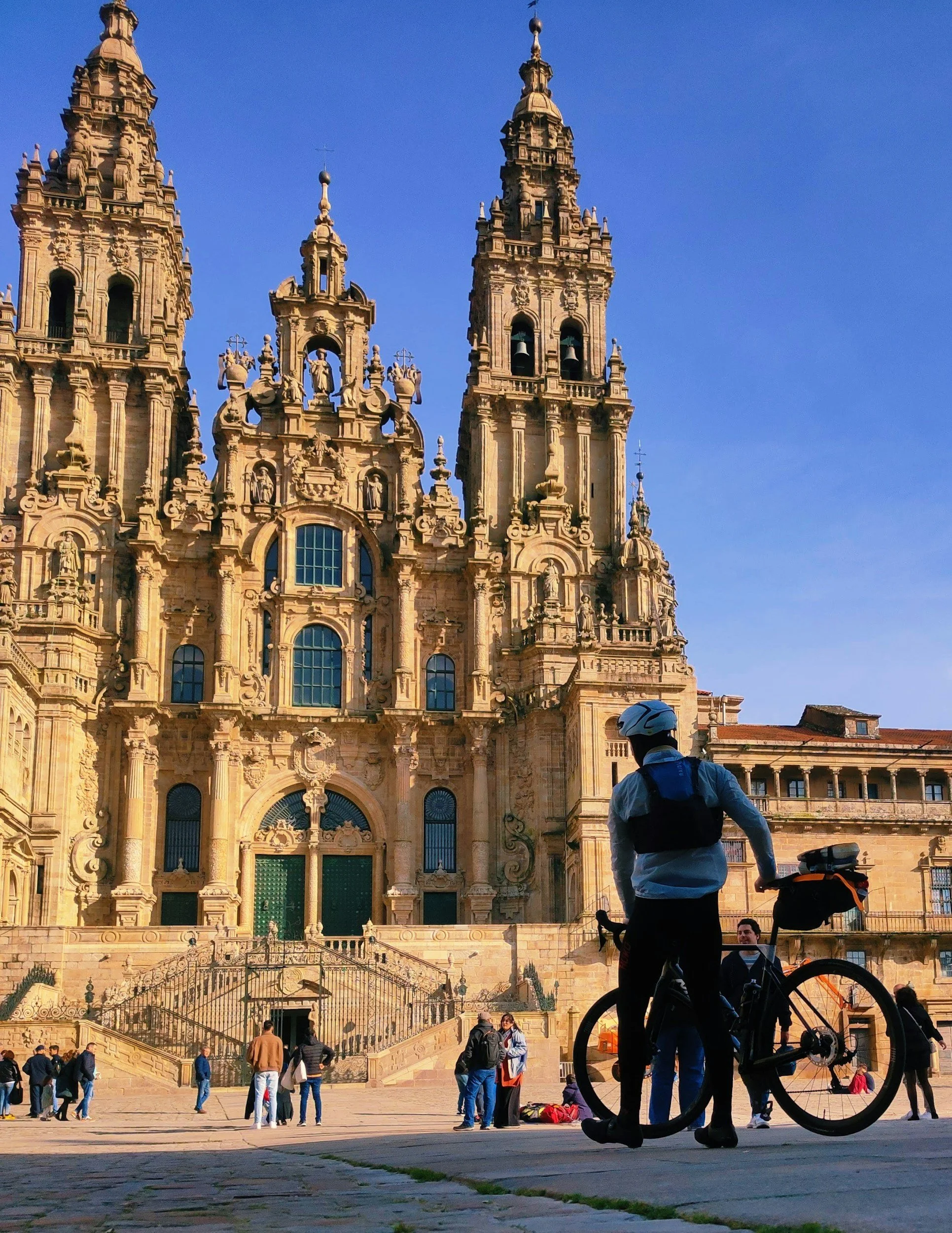 Vista de la catedral en una plaza concurrida. Personas caminan y conversan, un ciclista con casco y mochila está en primer plano, y varios turistas y locales disfrutan del día soleado.