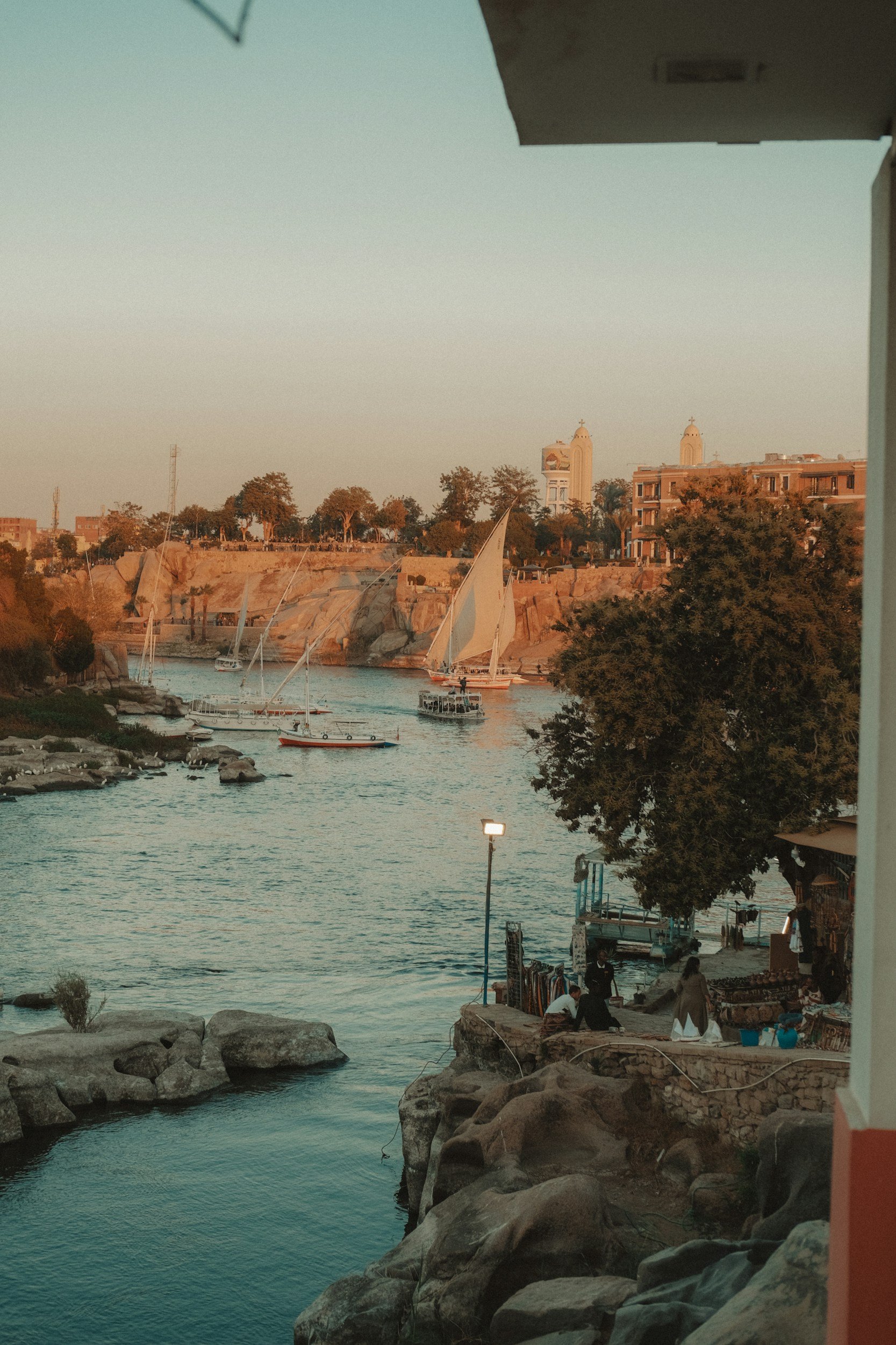 Vista de un río con barcos sailboat en el rio Nilo y una orilla con rocas y árboles, edificios al fondo con torres y vegetación, durante atardecer