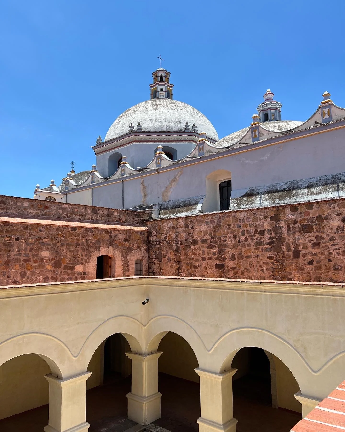 Iglesia con cúpula blanca y detalles en colores claros, con un muro de piedra en primer plano y un cielo azul despejado.