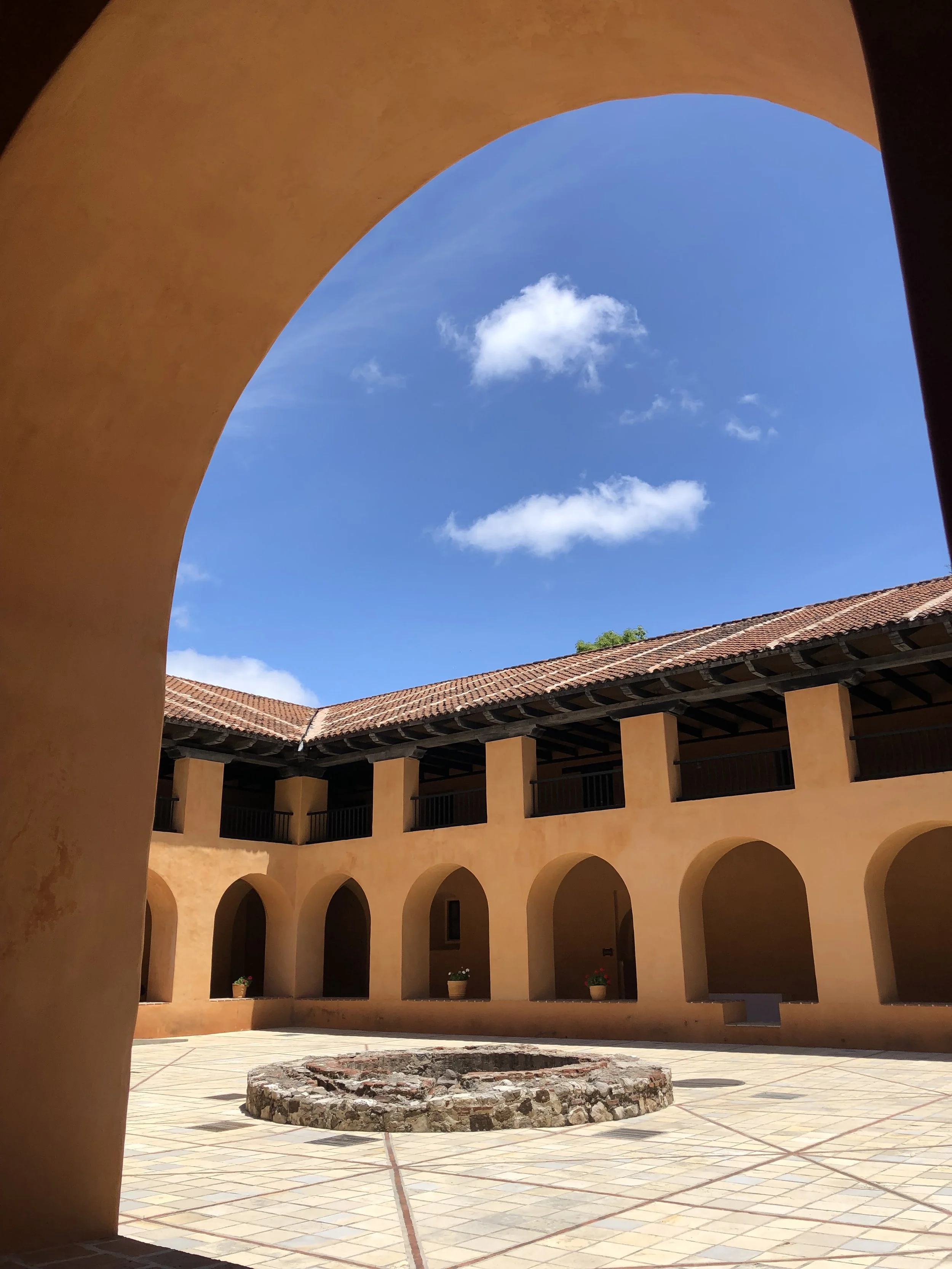 Vista del patio central de una edificación colonial con arcos, paredes de color cálido, un pozo de piedra en el centro y un cielo despejado con pocas nubes.