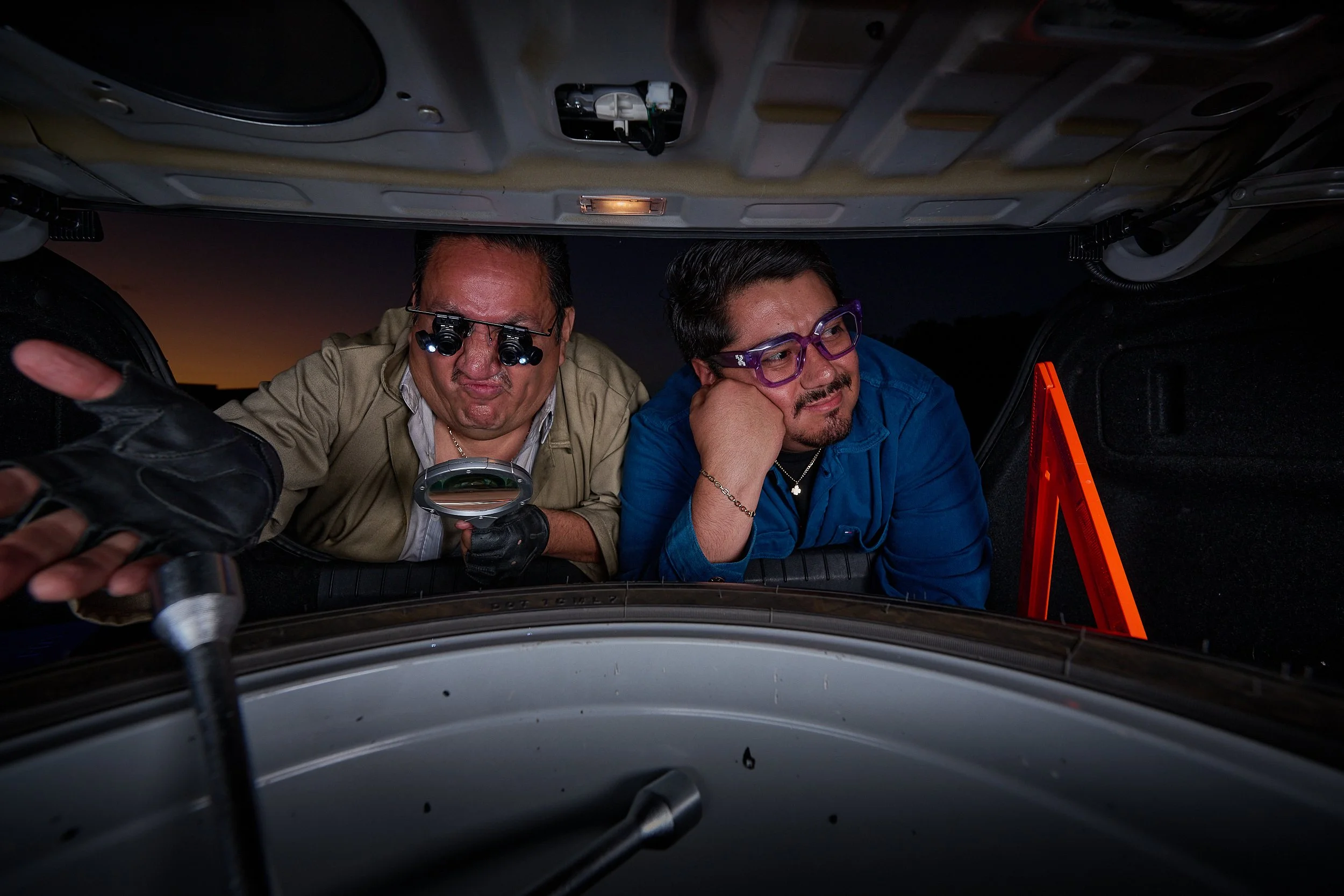 Two men lying underneath a vehicle, inspecting the underside. One man is wearing glasses with a headlamp, holding a flashlight. The other man is resting his head on his hand and looking up at the vehicle. It appears to be evening or night outside.