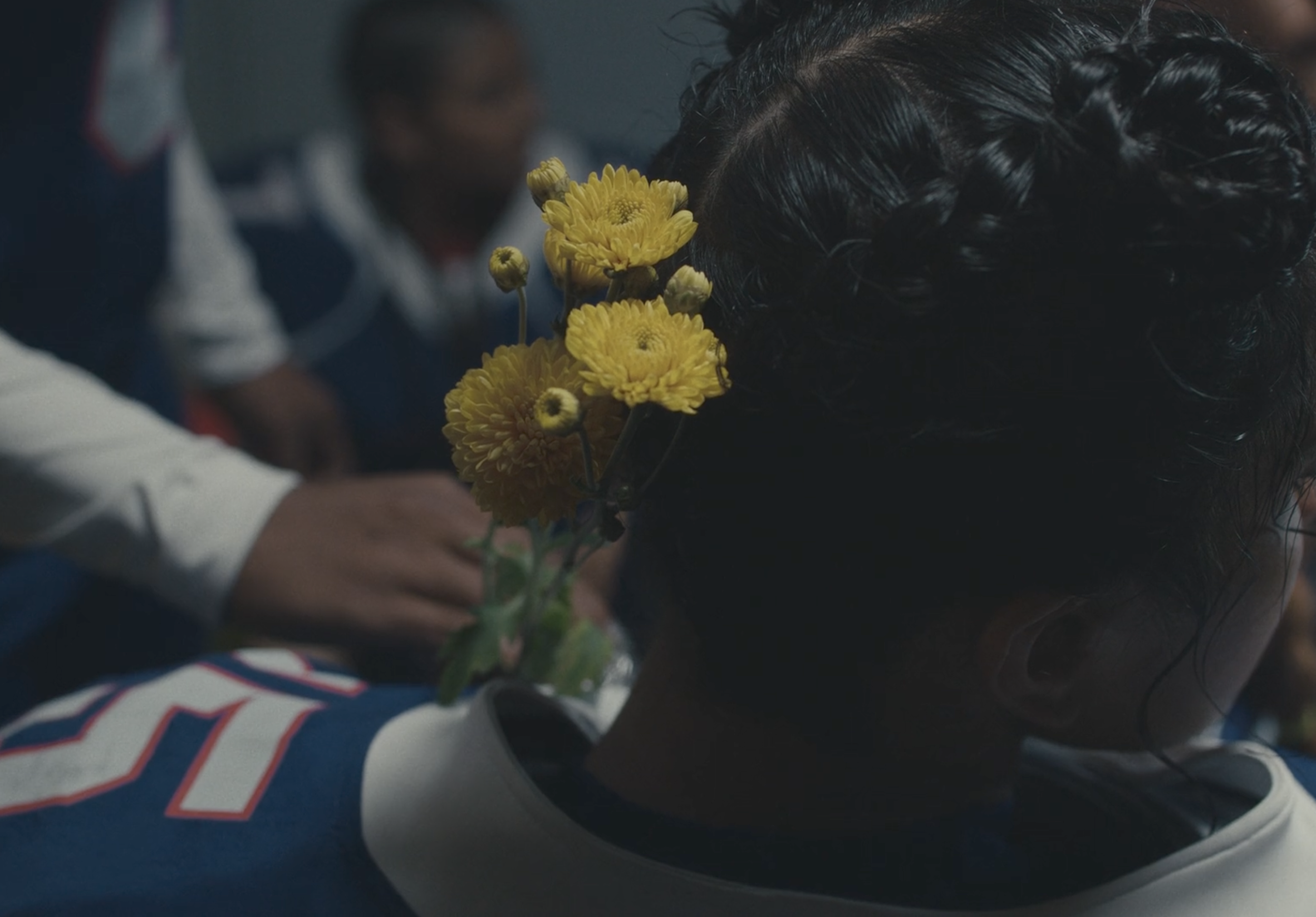 Close-up of a person with dark curly hair with yellow chrysanthemum flowers placed near the hair, possibly during a celebration or ritual. The background is out of focus with two other individuals, one wearing a striped shirt.