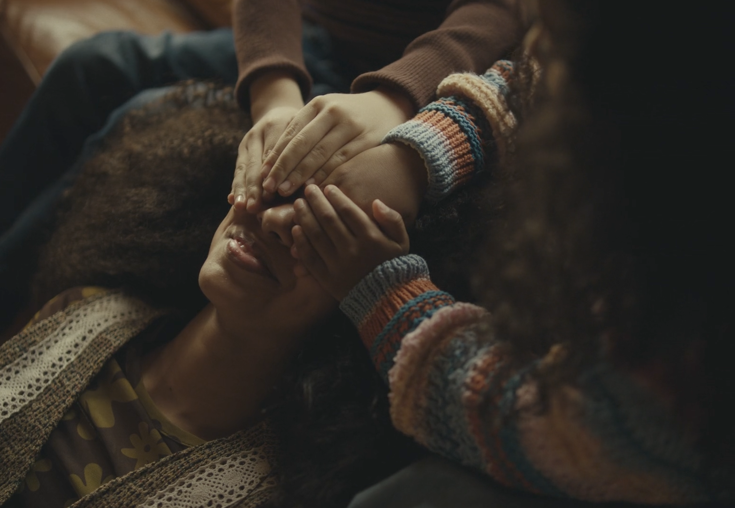 Two people holding hands, one person covering the other's face, in a warm setting with cozy clothing.