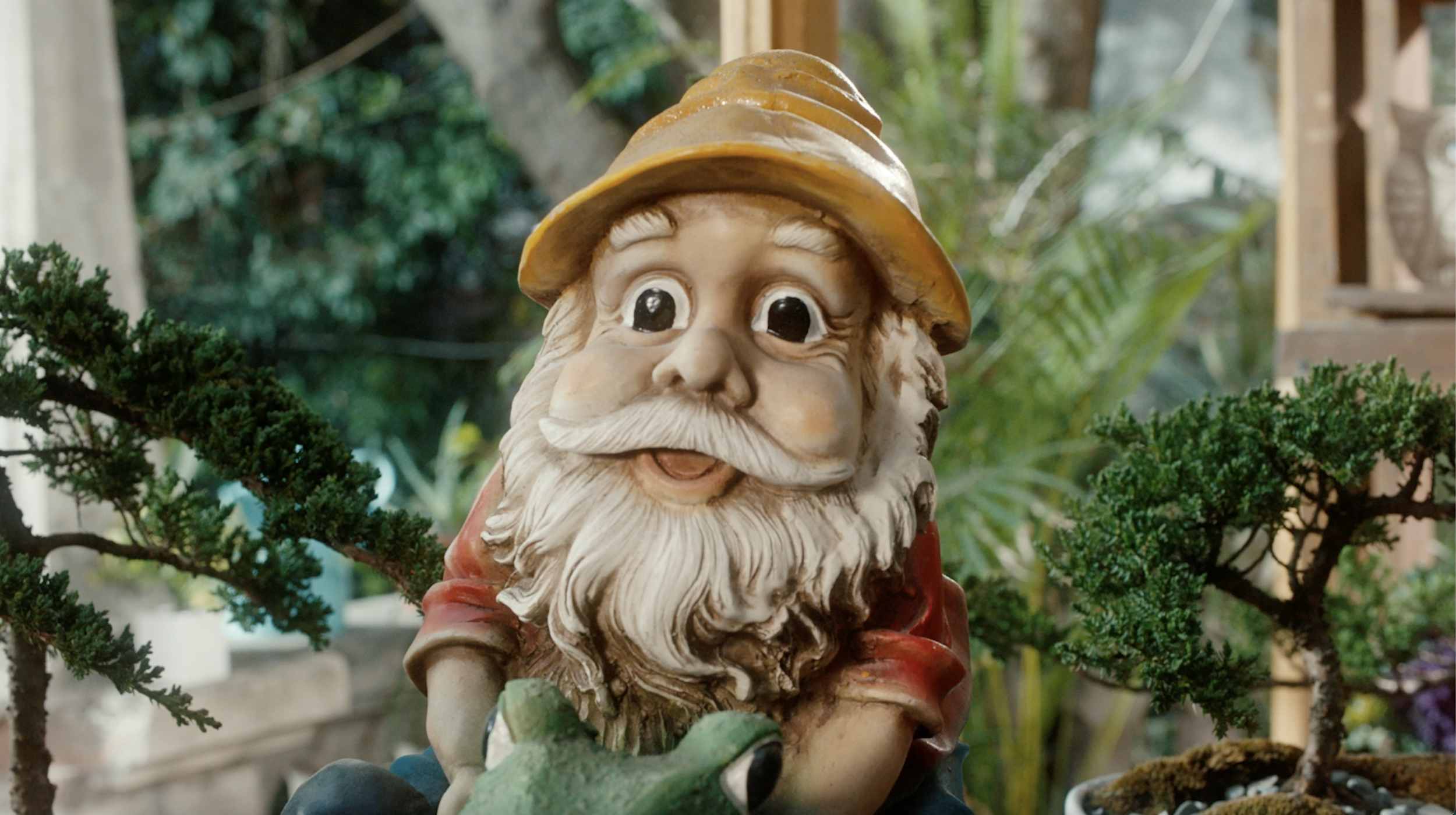 Figura de Santa Claus con barba blanca, sombrero amarillo, y expresión alegre, rodeada de plantas verdes en un entorno natural.