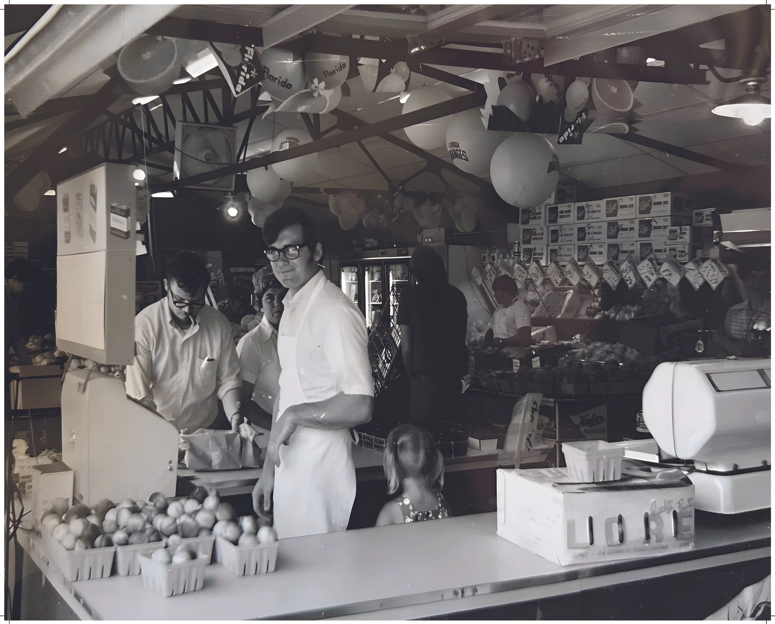 Black and white photo of Brennan's Market in Monroe. Skip Brennan is pictured in the produce section with tomatoes and other vegetables, surrounded by balloons and empty boxes