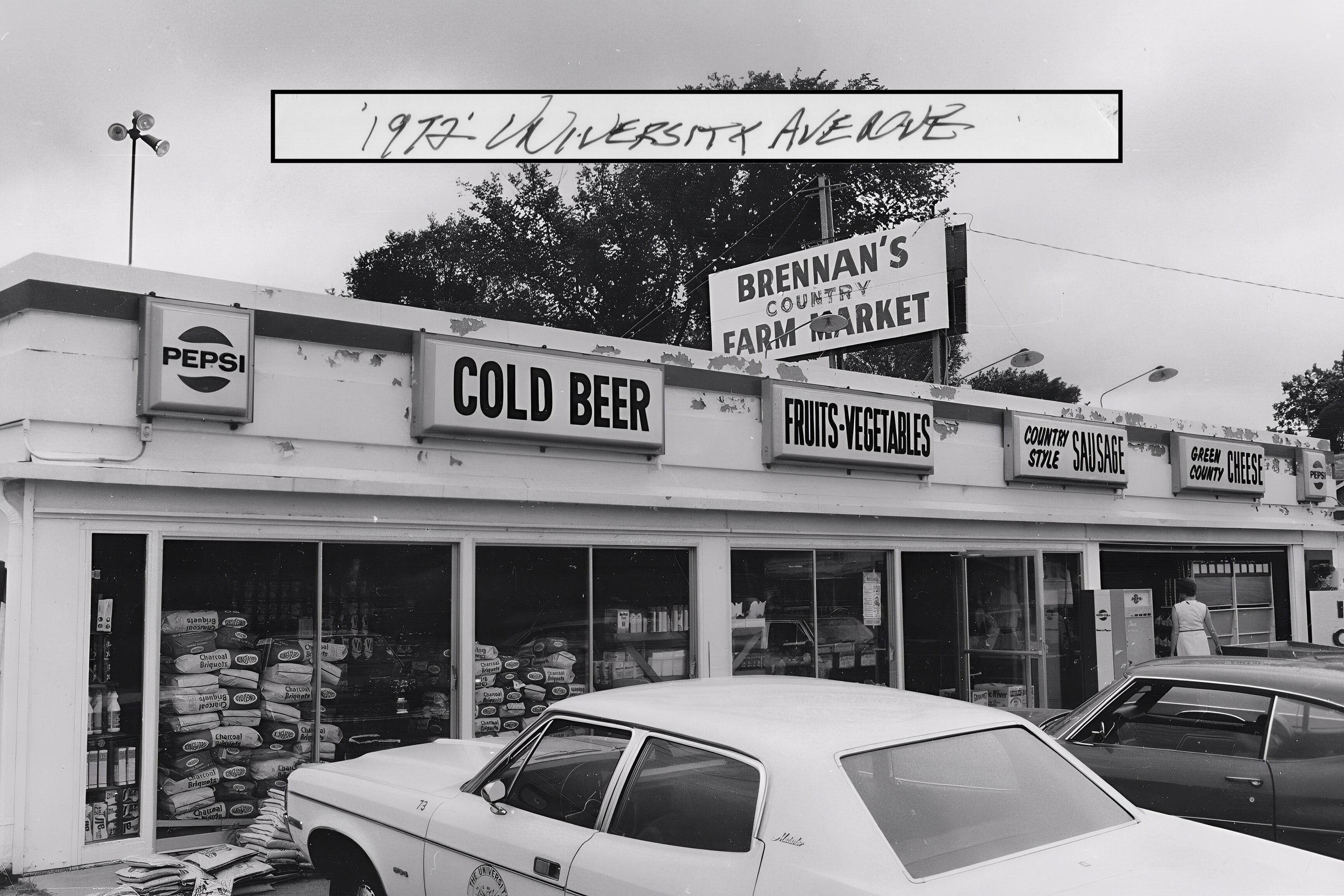 Black and white from 1972 of the old Brennan's store on Univeristy Avenue with signs advertising Pepsi, cold beer, fruits and vegetables, country style sausage, and County cheese. There are cars parked in front and a person walking nearby.