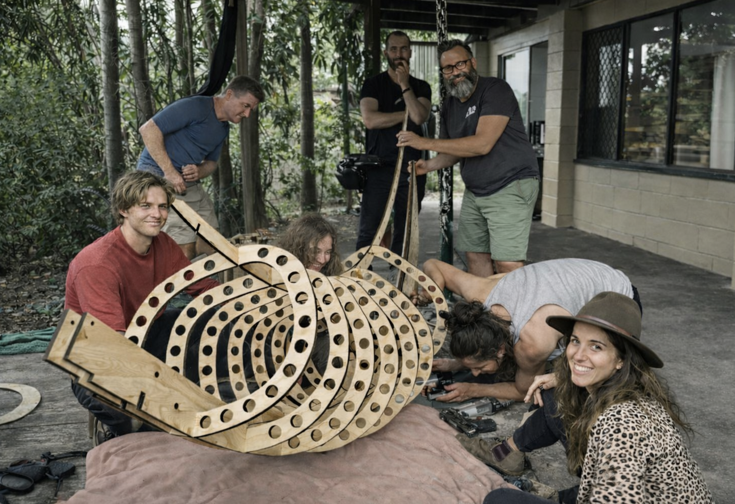Group of six people working together on a wooden snake sculpture outside on a patio, surrounded by trees and a building with large windows.