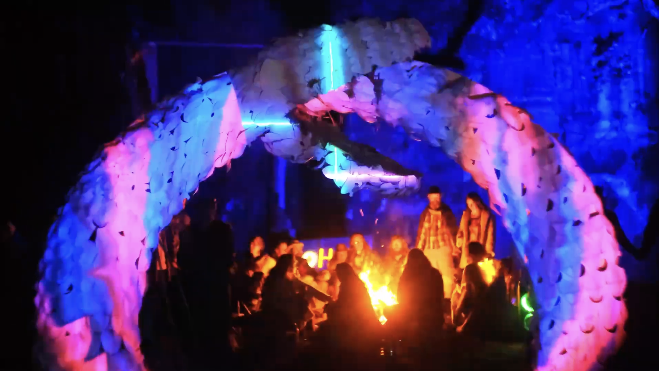 People gathered around a campfire under a decorated archway with colorful lights at night.